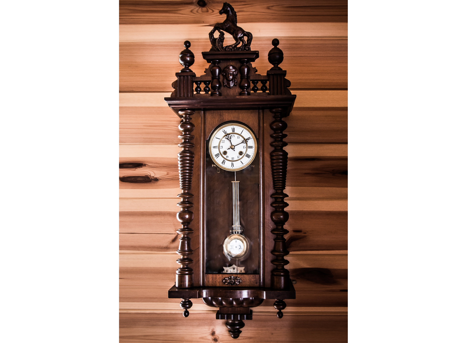 Zdjęcie zegara z wahadłem. Obudowa zegara z drewna, kunsztownie wykonana. Szczyt zwieńczony jest koniem stojącym na tylnych nogach. Na tarczy zegara cyfry rzymskie. Wskazówki wskazują godzinę jedenastą dziesięć. Poniżej tarczy wahadło z napędem sprężynowym. Soczewka, a więc okrągły ciężarek, zawieszona jest na pręcie z układem kompensującym niepożądany wpływ warunków zewnętrznych. Układ kompensujący to kilka prętów równoległych do głównego pręta, połączonych u szczytu i u dołu. Za zegarem drewniana powierzchnia.