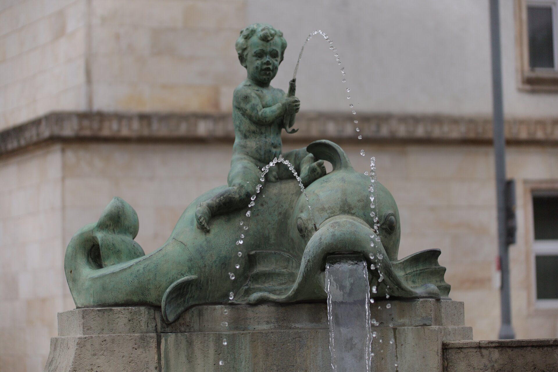 Ilustracja przedstawia fontannę, w której znajduje się rzeźba chłopczyka siedzącego na dużej rybie. Chłopczyk jest nagi, ma krótkie kręcone włosy. W rękach chłopczyk trzyma małą rybkę, z której ust wydobywa się woda, z ust dużej ryby również wylatuję woda. Rzeźba znajduję się na kamiennym podwyższeniu.