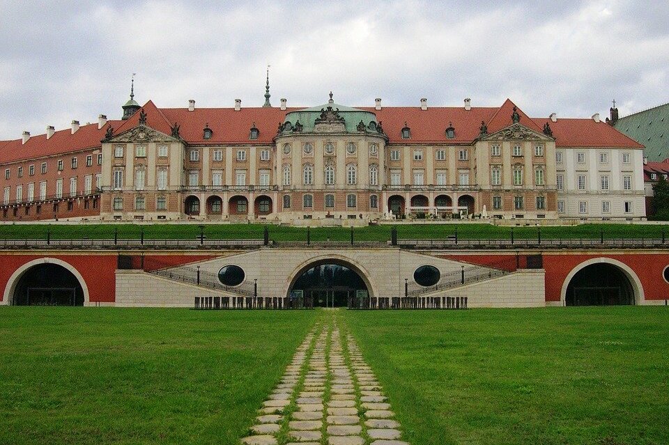 Zdjęcie przedstawia Zamek Królewski w Warszawie. Jest to ogromny kompleks budynków. Na pierwszym planie widać ścianę z muralem imitującym dziedziniec pałacu. Po środku ściany jest wejście ze szklanymi drzwiami na prawdziwy dziedziniec zamku.