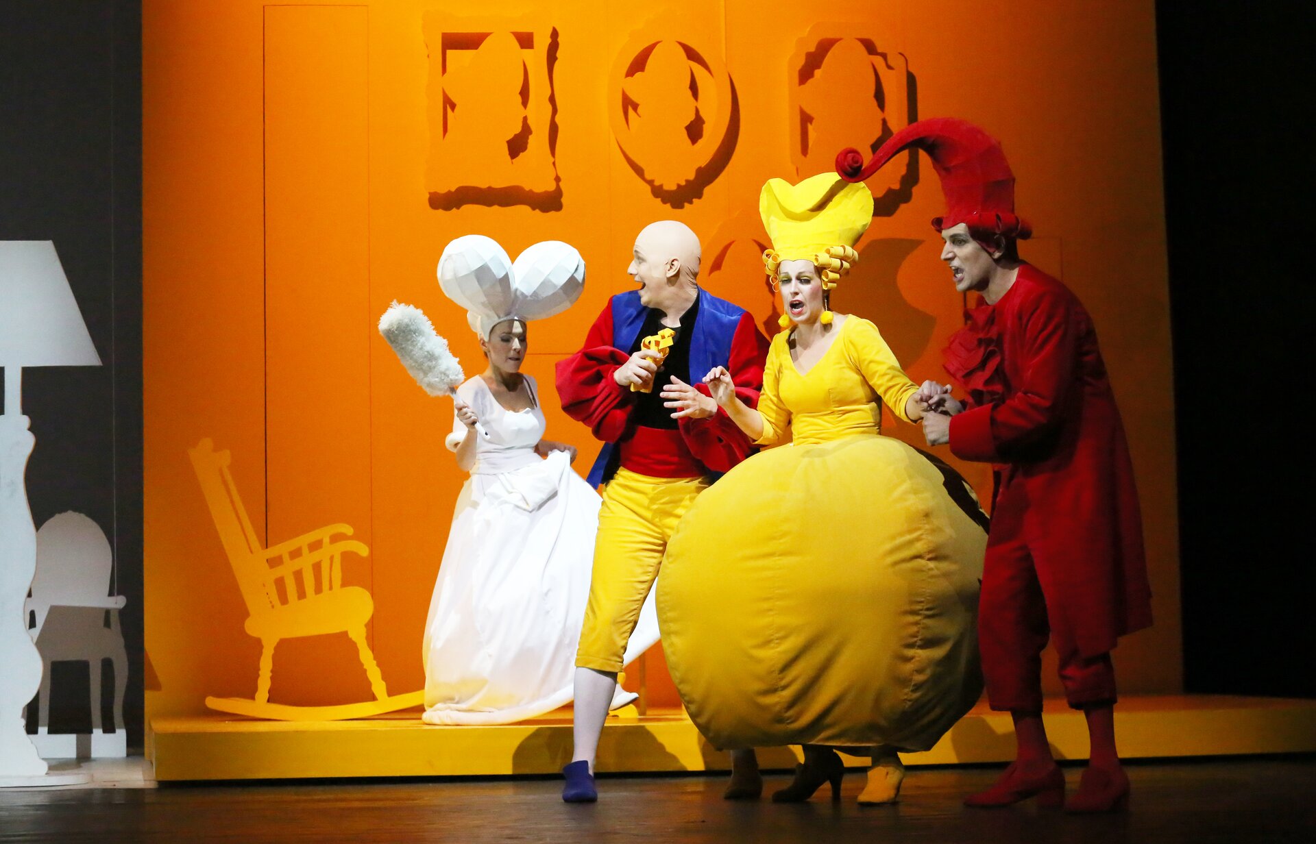 Na zdjęciu widoczne są cztery osoby: kolejno od lewej strony: kobieta ubrana w białą, długą suknię, na głowie posiada duży biały element/czapkę, trzyma także białą miotełkę do kurzu. Obok stoi mężczyzna ubrany w czarną bluzkę, żółte spodnie, białe rajstopy i niebieskie buty. Ma na sobie także niebieską narzutę/kurtkę z czerwonymi, bufiastymi rękawami – trzyma on żółty element. Druga kobieta stojąca obok ma na sobie żółtą bluzkę z długim rękawem oraz żółtą spódnicę, która przypomina kulę, a także żółte buty na obcasie. W uszach ma żółte kolczyki w kształcie kul, a na głowie żółtą, dużą czapkę. Drugi mężczyzna (trzymający ją za rękę) posiada cały czerwony strój – spodnie, długą bluzkę z dekoracyjnym żabotem, rajstopy jak i buty. Osoby te znajdują się na scenie o drewnianej podłodze. Sceneria to pokój o pomarańczowych ścianach i podłodze. Atrapy obrazów wiszą na ścianie i także są pomarańczowe (jak i bujające się krzesło). Po lewej stronie ilustracji widoczna jest biała atrapa lampy i krzesła.