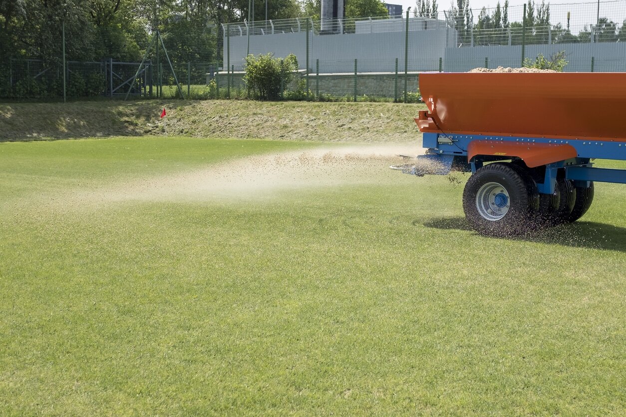 Zdjęcie przedstawia piaskowanie torfu. Widać pojazd rozrzucający piasek po trawniku.
