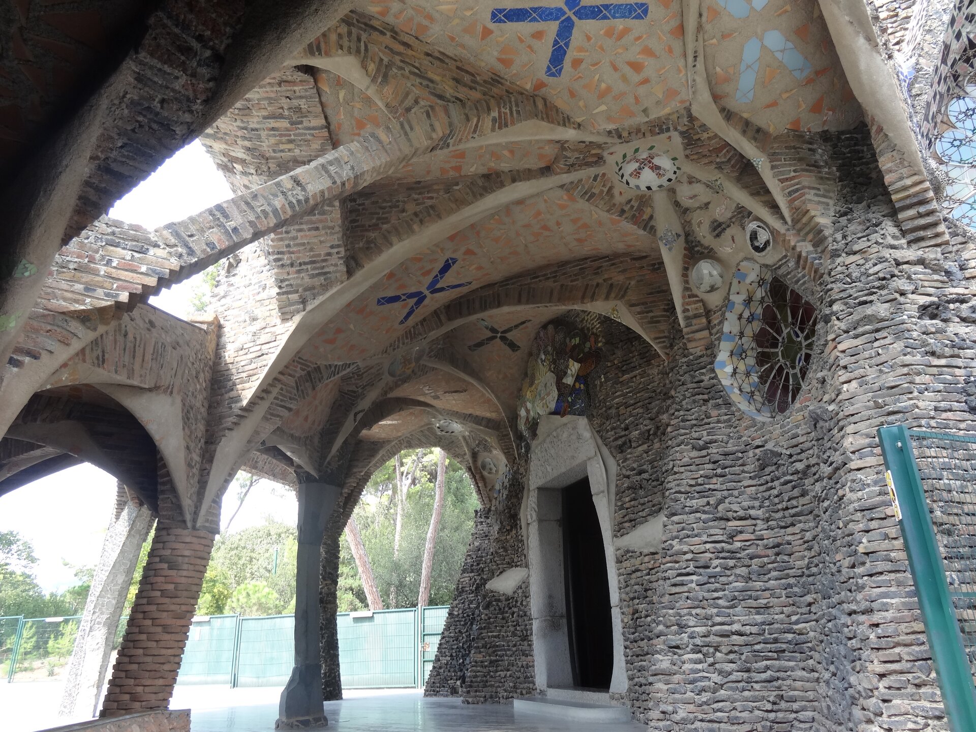 Ilustracja o kształcie poziomego prostokąta przedstawia portyk Krypty w kolonii Guell, Santa Coloma de Cervelló, zaprojektowany przez Gaudiego. Obiekt ma szaro-brązową barwę, Kolumny zewnętrzne posiadają  kształt rosnących pni wyrastających z ziemi i połączonych z bryłą łukami o zróżnicowanych kształtach. Do wewnątrz prowadzi niewielkie wejście otoczone białym tynkiem. W ściennie znajduje się okrągłe okno dekorowane na styl gotyckiej rozety.