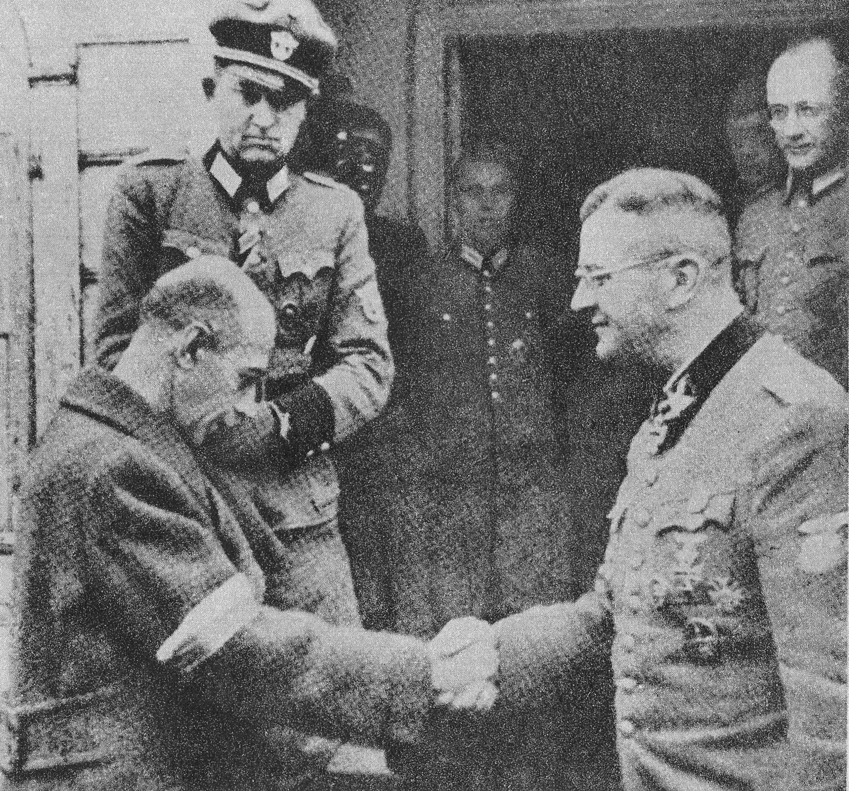 Kliknij, aby powiększyć Zdjęcie przedstawia dwóch mężczyzn, którzy podają sobie dłoń. Mężczyzna po lewej ma opuszczoną głowę. Ubrany jest w płaszcz z opaską. Mężczyzna po prawej nosi niemiecki mundur z odznaczeniami i orderami na piersi. Patrzy na swojego rozmówcę i się uśmiecha. W tle kilku niemieckich oficerów i ściana budynku.