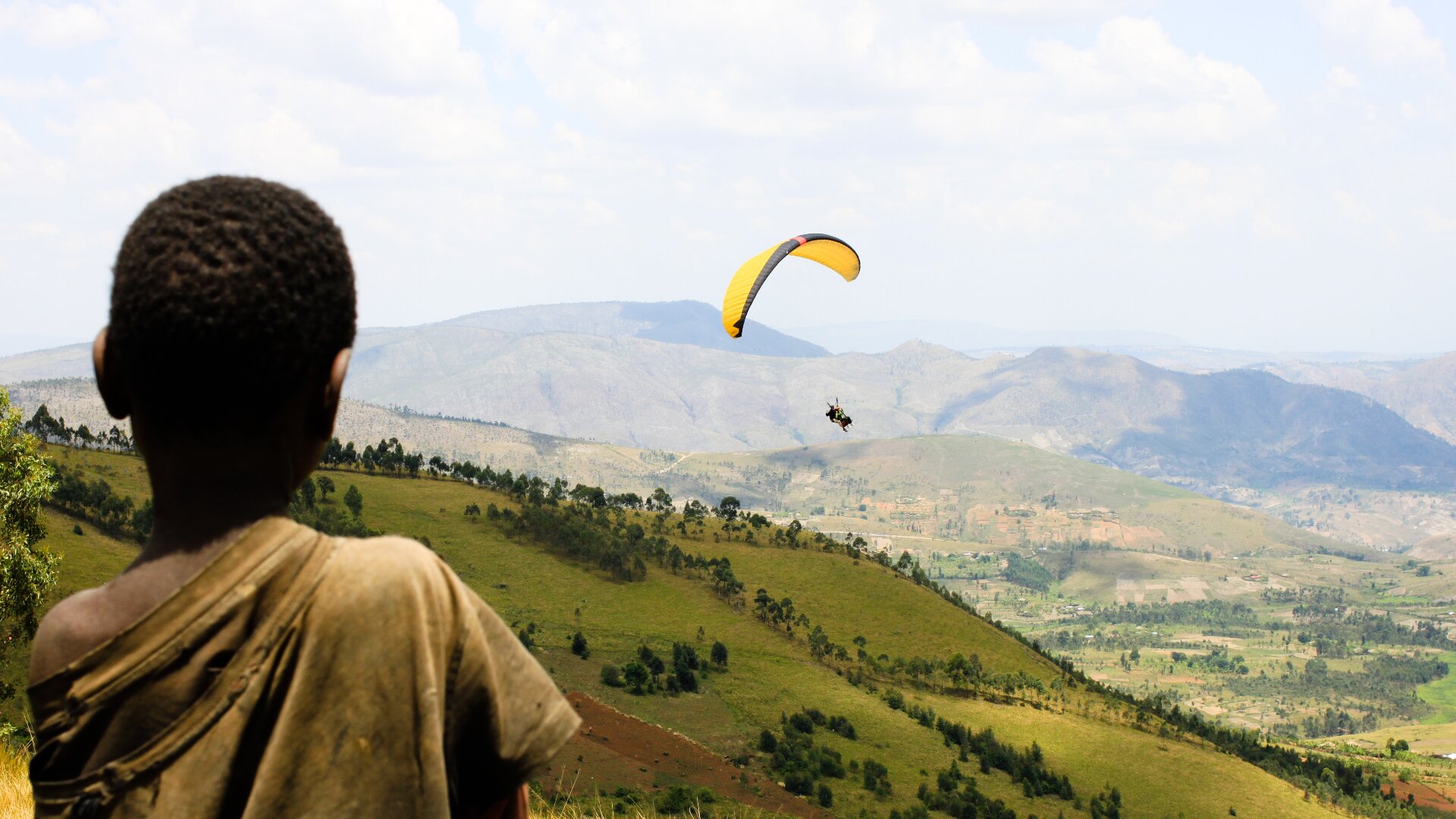 Zdjęcie przedstawia Burundi. Na pierwszym planie tyłem stoi czarnoskóry chłopiec. Za nim są zbocza porośnięte trawą i drzewami. Nad nimi lata paralotniarz. W tle są góry.