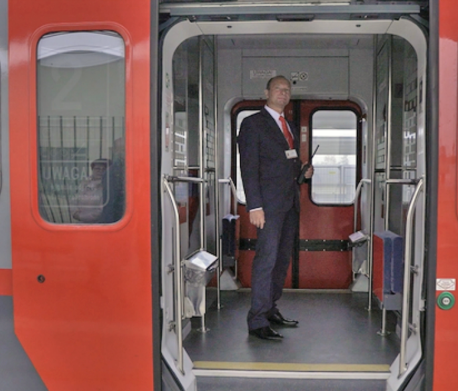 Zdjęcie przedstawia kierownika pociągu. Na zdjęciu widać mężczyznę przechodzącego wzdłuż wagonu pomiędzy fotelami w wagonie pasażerskim bezprzedziałowym. Na ramieniu niesie on torbę.