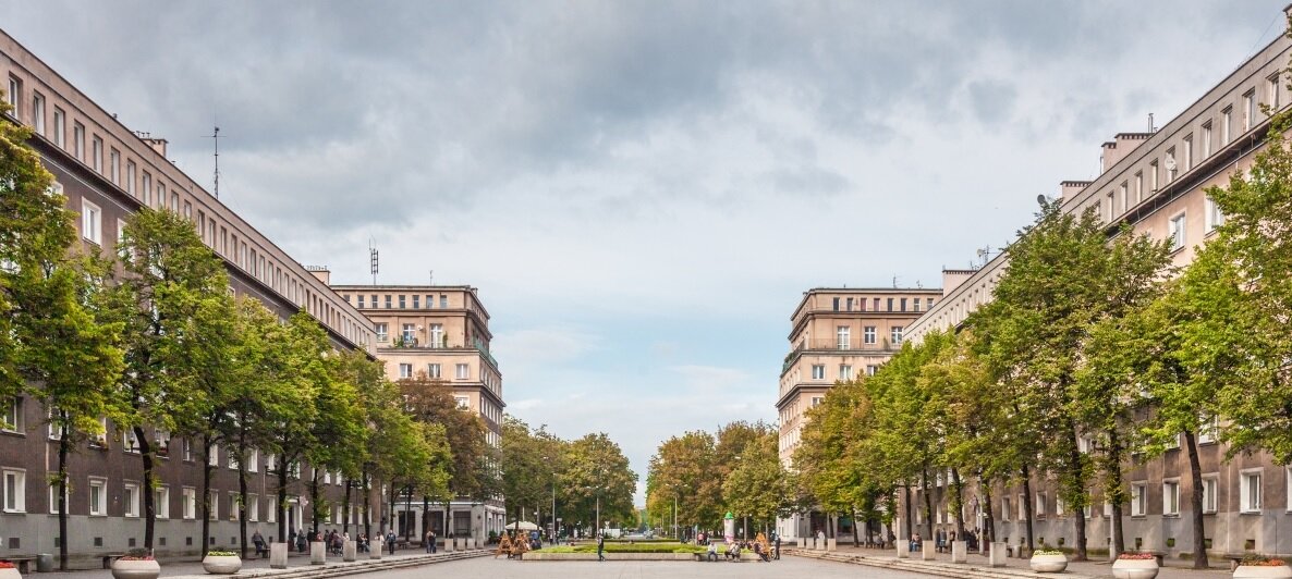 Zdjęcie przedstawia szeroką aleję z rzędami drzew w mieście. Po jej lewej i prawej stronie stoją identyczne budynki. Mają sześć pięter, na ich końcu jest nieco wyższa część. Budynki mają prostą konstrukcję w kształcie prostokątów.