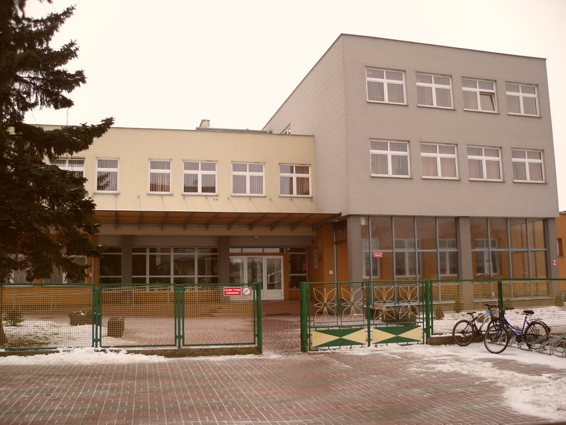 Ilustracja przedstawia budynek trzykondygnacyjny w kolorze szarym. Drzwi i okna występują w kolorze białym. Plac przed budynkiem wyłożony jest kostką brukową. Budynek ogrodzony jest metalowym płotem wykonanym z siatki w kolorze zielonym.