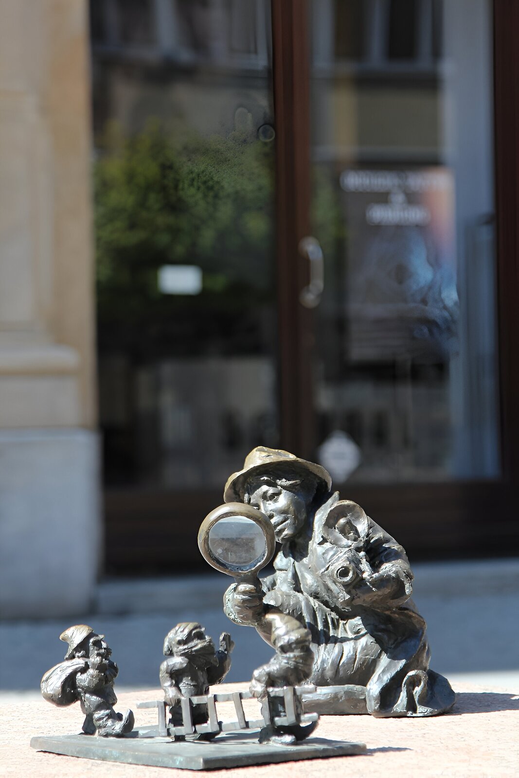 Zdjęcie przedstawia rzeźbę trzech krasnali obserwowanych przez małego chłopca za pomocą lupy. Chłopiec na głowie ma kapelusz, a w lewej ręce trzyma aparat fotograficzny. Krasnale idą przed siebie jeden za drugim. Pierwsze dwa niosą drabinę, a trzeci niesie drugi worek.