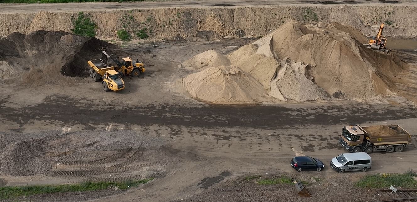 Ilustracja przedstawia proces korytowania. Widoczne są tu koparki oraz wywrotki podczas pracy. Na środku zdjęcia znajduje się hałda usypana z urobku.