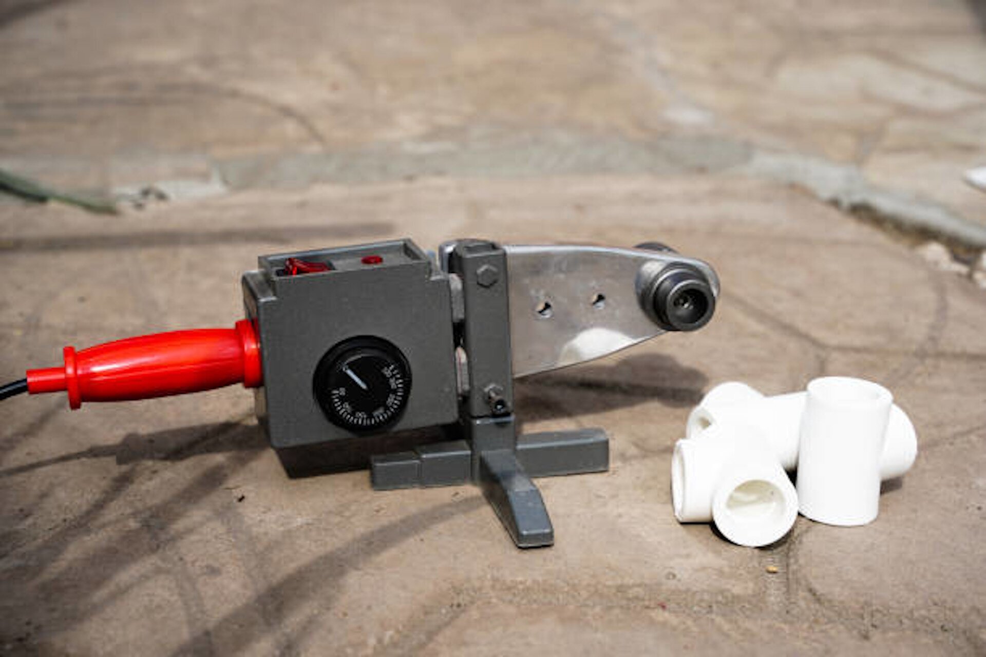 Zdjęcie przedstawia zgrzewarkę do rur. Urządzenie jest  małe i wyposażone w kabel zasilający. Korpus zgrzewarki wykonany jest z wytrzymałej stali, a na jego powierzchni znajdują się przycisk włączający oraz pokrętło do regulacji temperatury nagrzewania. Na końcu zgrzewarki znajdują się dwa pierścienie, które służą do umocowania rur w czasie zgrzewania. Obok zgrzewarki widoczne są kawałki rur, które są łączone za pomocą tego narzędzia.