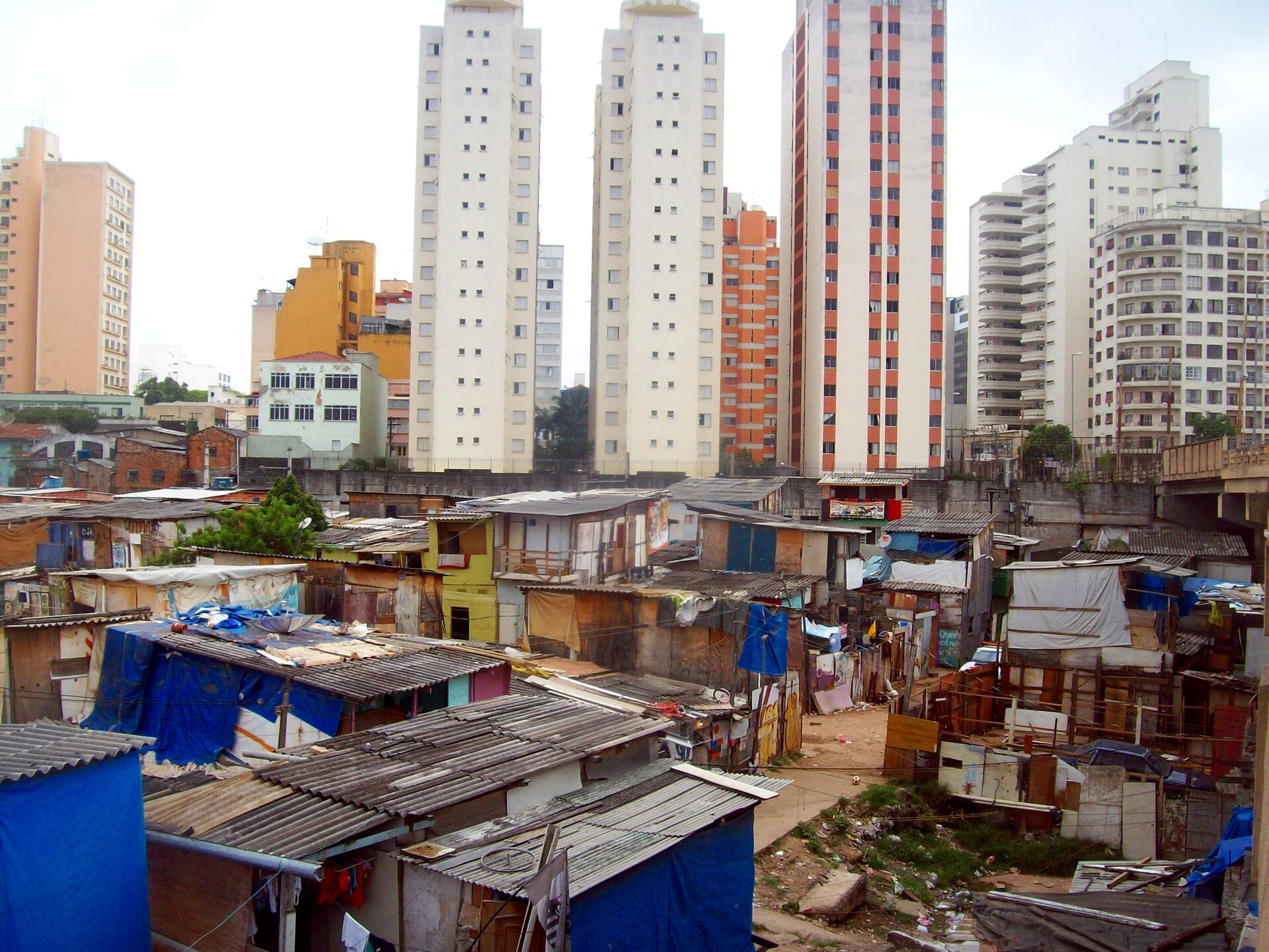 Na zdjęciu widoczna jest dzielnica mieszkaniowa w Sao Paulo. Na pierwszym planie znajduje się favela z prowizorycznymi domami. Domy zbudowane z najtańszych materiałów: tektury, dykty, blachy, desek. W tle widoczne są nowoczesne wieżowce na tle białego nieba.