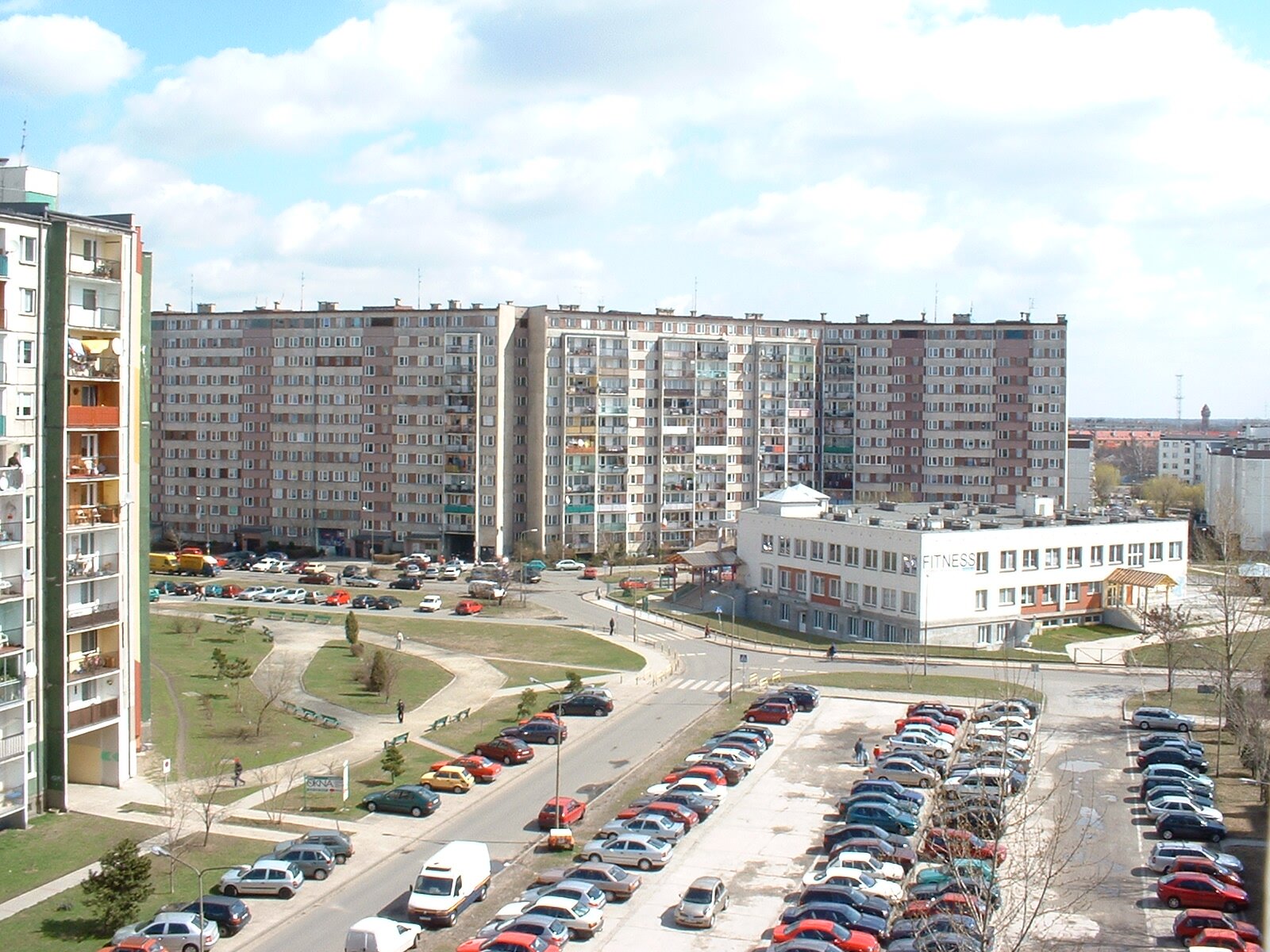 Zdjęcie przedstawia parking i samochody. Wokół parkingu stoją wieżowce i dwupiętrowy budynek użyteczności publicznej. 
