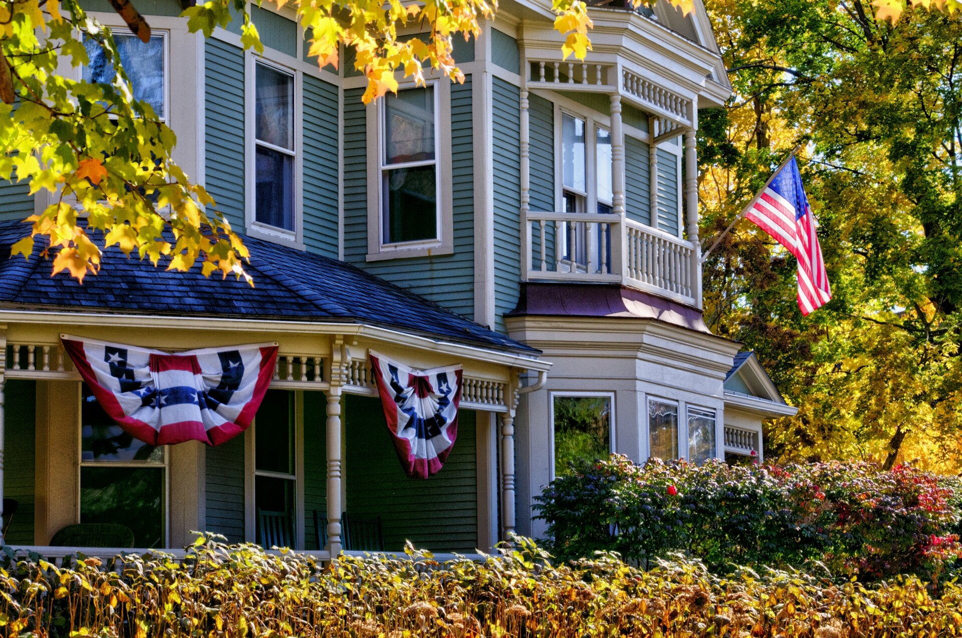 Zdjęcie przedstawia fasadę drewnianego domu w kolorze niebieskim, z białymi detalami architektonicznymi. Fasadę widać lekko z ukosa. Po lewej stronie widoczny jest ganek udekorowany dwoma, upiętymi w wachlarze, amerykańskimi flagami tworzącymi kotyliony. Pośrodku, na niewielkim balkonie, widoczna jest powiewająca amerykańska flaga. Całość otoczona jest drzewami i krzewami z żółtymi, pomarańczowymi i brązowymi liśćmi wskazującymi na porę jesienną.