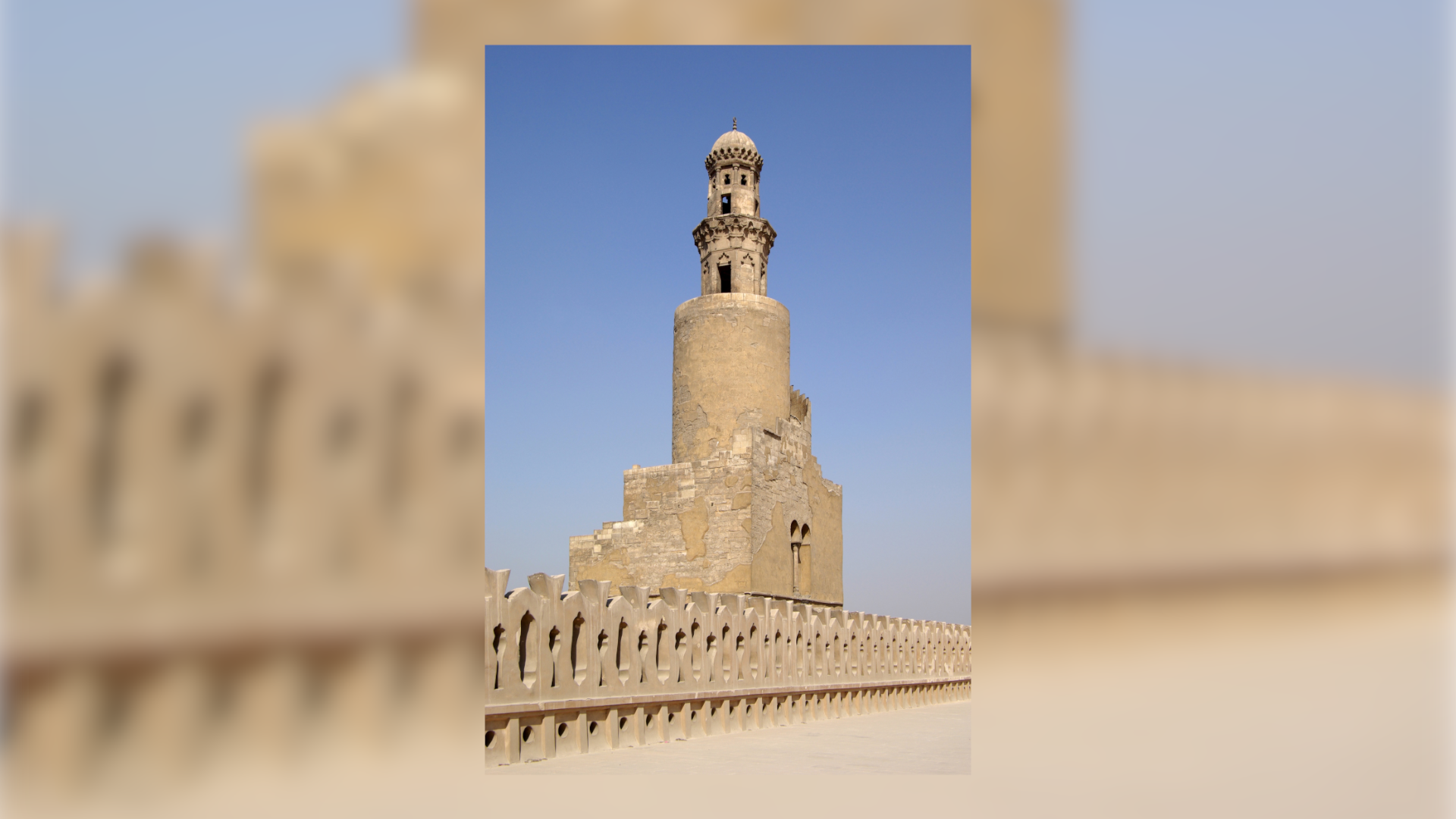 Na zdjęciu znajduje się lekko zniszczony kamienny minaret. Jest to masywna wysoka budowla. Jest otoczony niskim ozdobnym murem. 