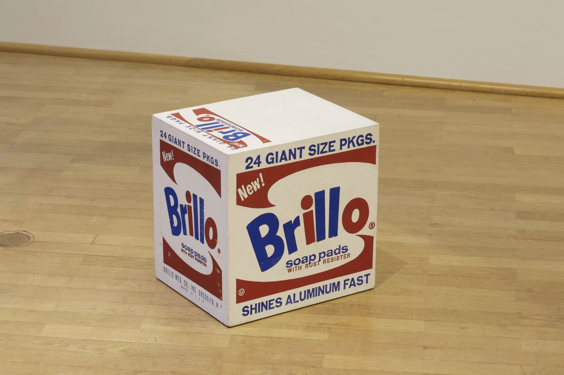 Ilustracja przedstawia fotografię projektu pudełka „Brillo Box”, sporządzonego przez Andy'ego Warhola. Ukazuje ujęcie perspektywiczne pudełka z widoczną ścianą górną i krawędzią boczną. Zawiera napis "Brillo" w niebiesko-czerwonym kolorze, symetrycznie przedstawiony, ale odwrócony motyw graficzny na dole i u góry w kolorze czerwonym oraz napisy. Wszystkie widoczne ściany boczne są takie same.