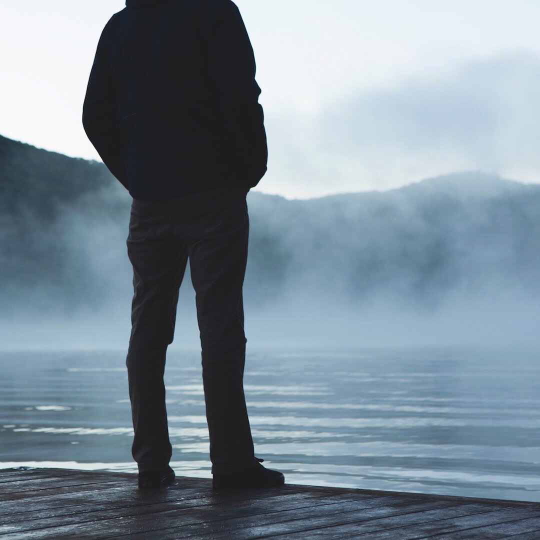 Zdjęcie przedstawia męską sylwetkę od barków do stóp w ciemnym ubraniu i butach. Stoi na drewnianym pomoście tyłem w stronę widza, przodem w stronę wody. Ręce ma w kieszeniach. Nad wodą unosi się mgła. W oddali widać góry.