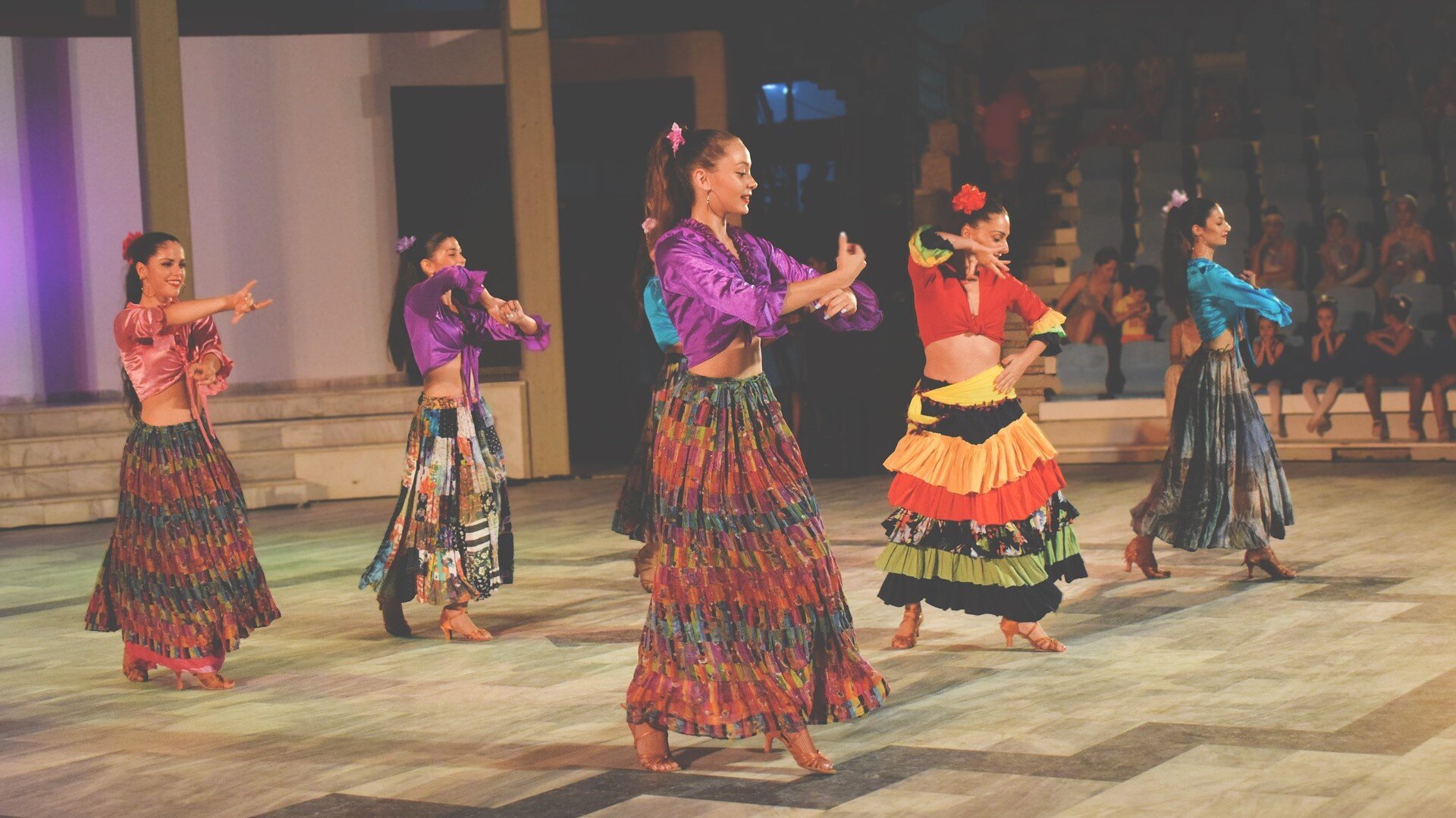 Zdjęcie przedstawia sześć kobiet tańczących flamenco na parkiecie. Ubrane są w długie kolorowe spódnice oraz kolorowe krótkie bluzki i buty na obcasie.