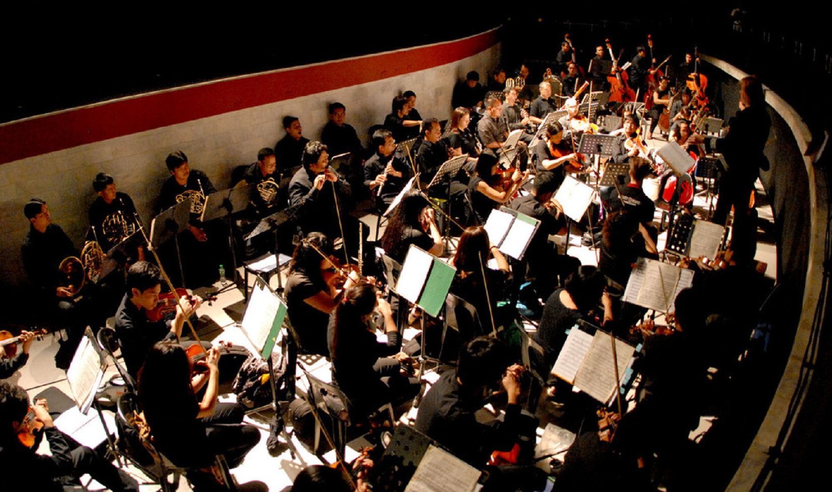 Zdjęcie przedstawia orkiestron, wypełniony grającą orkiestrą. Krzesła są ustawione jedne przy drugim. Muzycy mają rozłożone pulpity z nutami. Po prawej stronie stoi dyrygent.