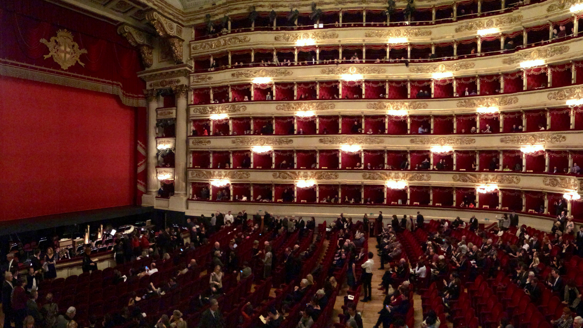 Zdjęcie przedstawia wnętrze teatru La Scala w Mediolanie. Na widowni widoczni są widzowie. Kurtyna jest opuszczona.