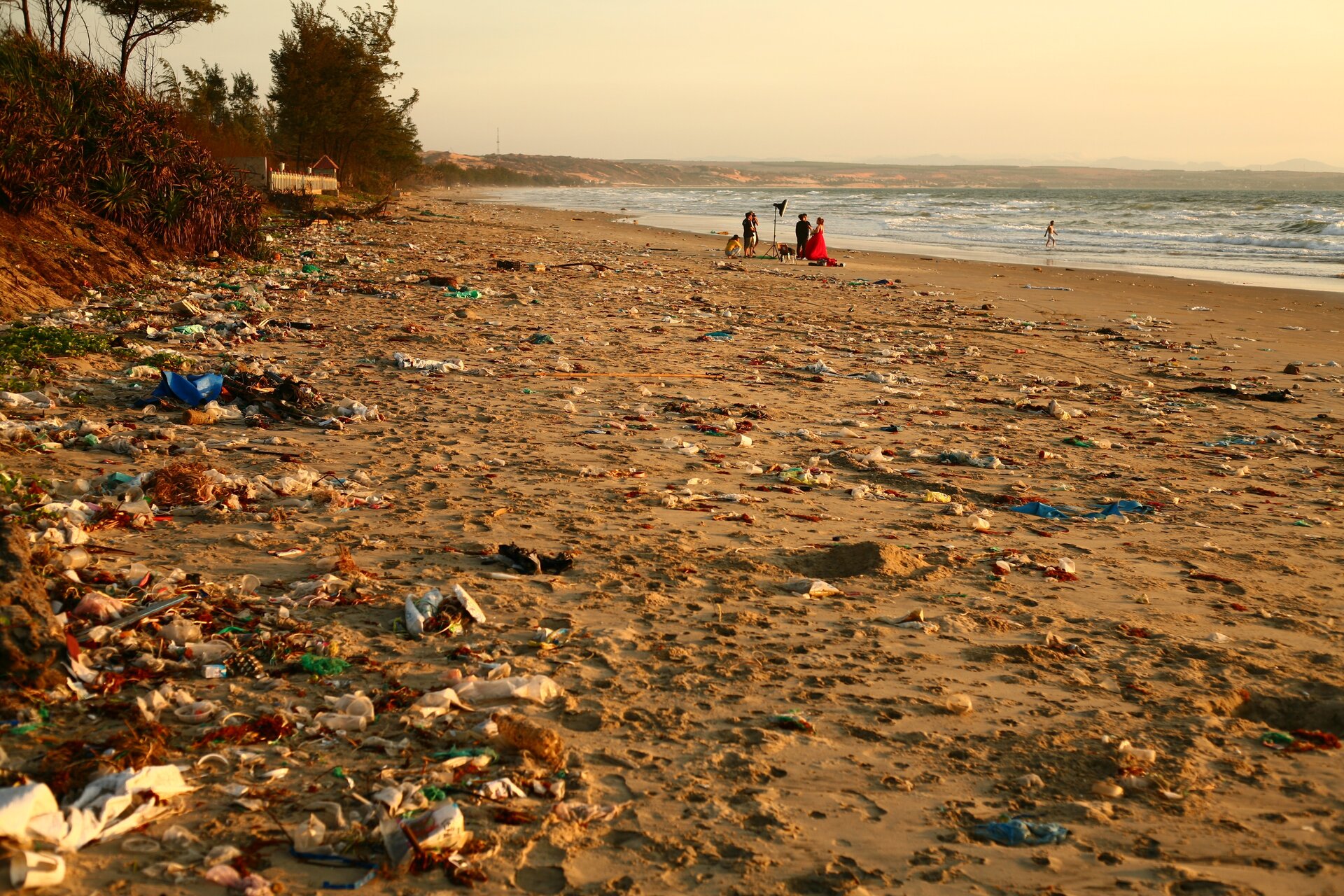 Ilustracja przedstawia plażę, na której znajduje się mnóstwo śmieci. Są to butelki, opakowania plastikowe i inne. W tle, przy brzegu oceanu widoczna jest grupa ludzi. Przy brzegu, w wodzie bawi się dziecko. 