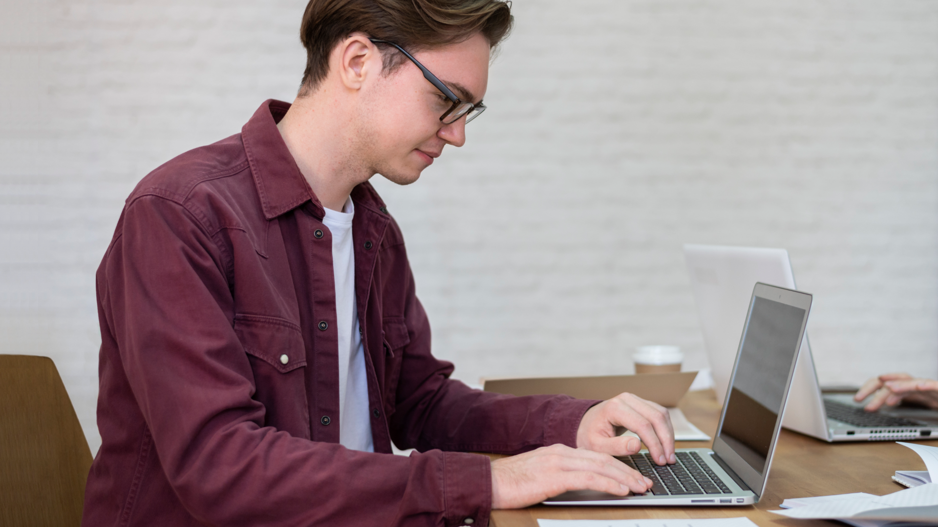 Zdjęcie przedstawia chłopaka w okularach siedzącego przy biurku przed laptopem. Chłopak siedzi bokiem do kadru.