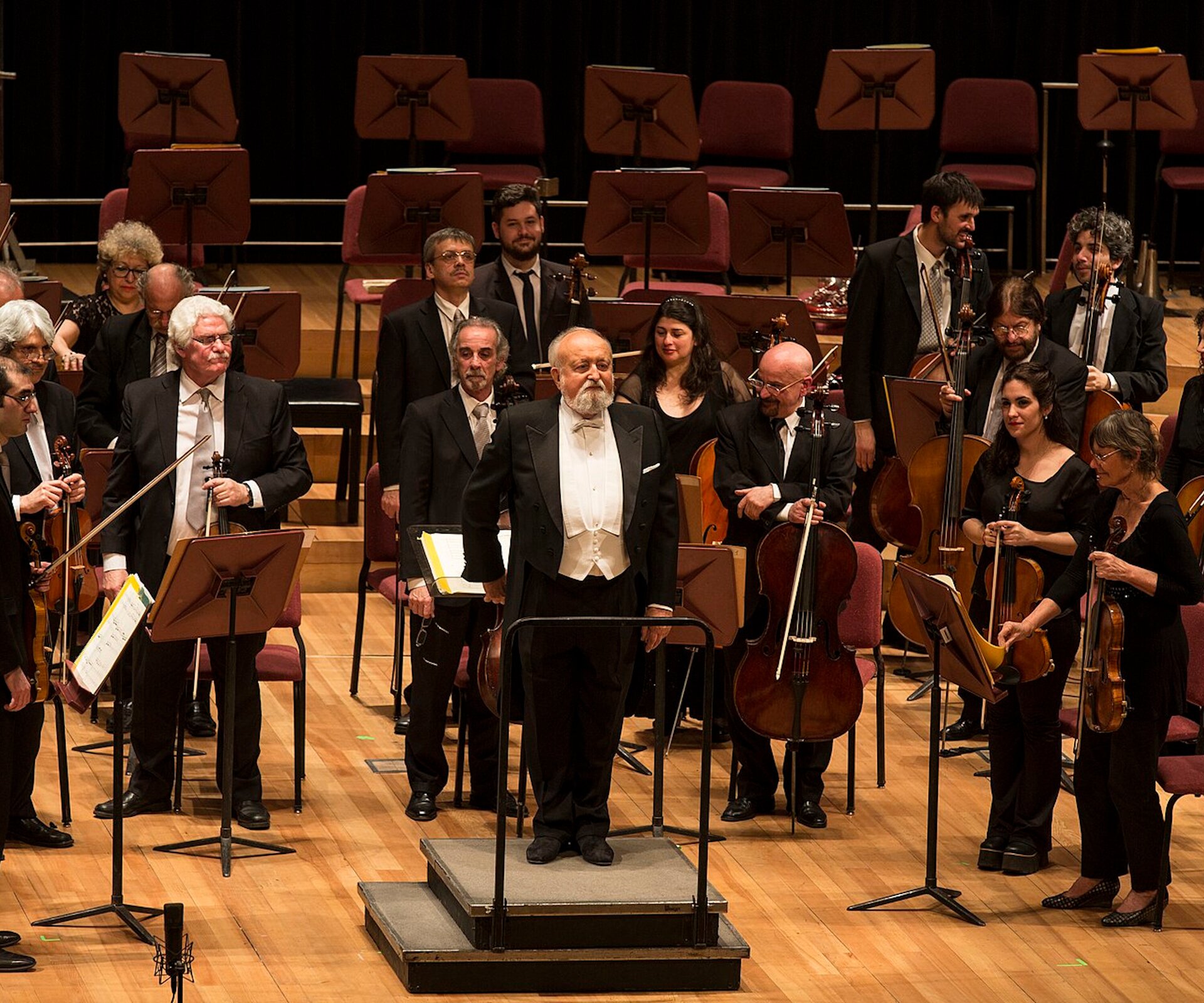 Na zdjęciu muzycy z instrumentami oraz dyrygent na sali koncertowej. Wszyscy stoją przodem do publiczności.