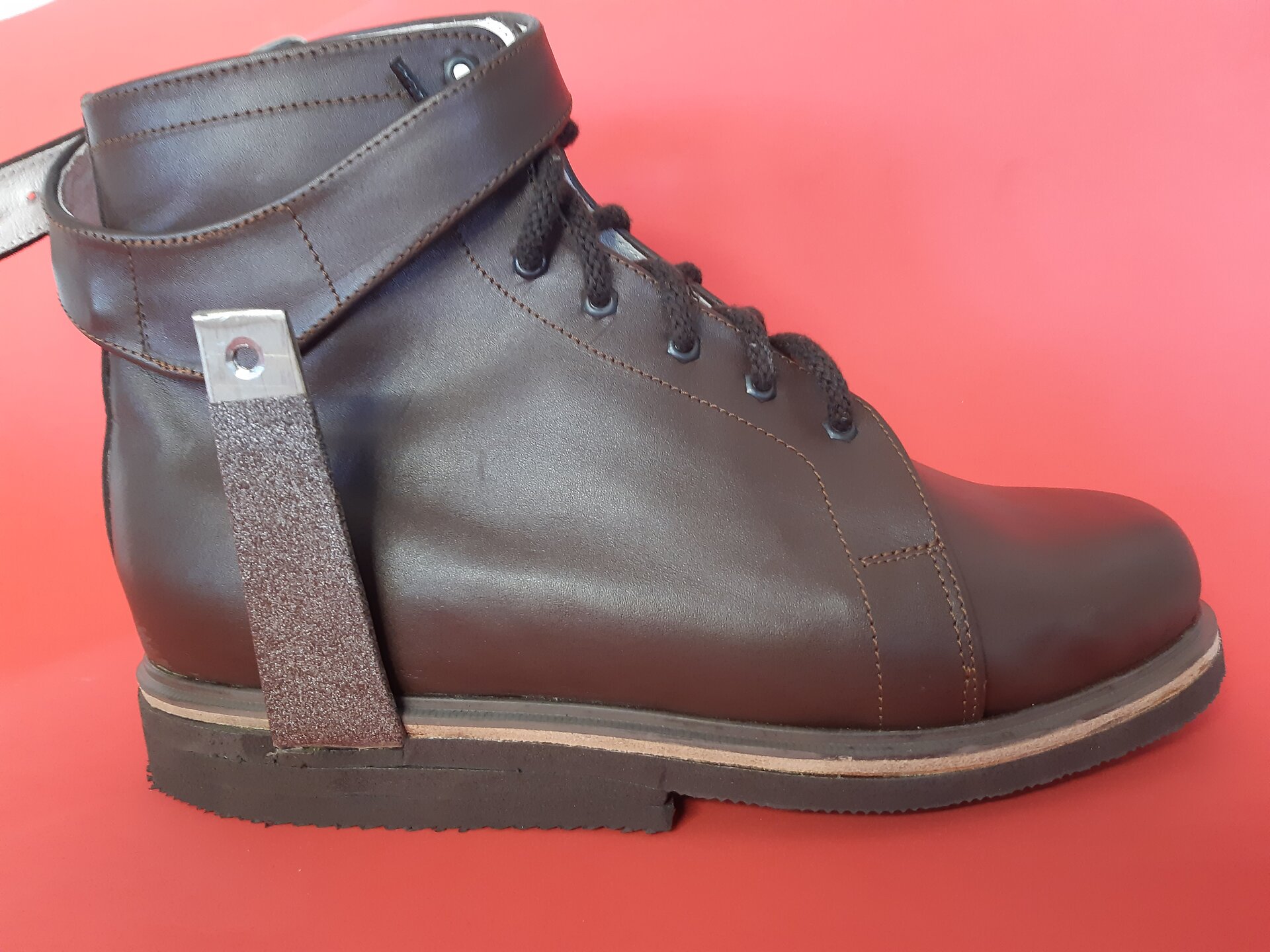 Zdjęcie przedstawia but ortopedyczny ze strzemieniem do ortezy, która przymocowana jest do zewnętrznej, tylnej części podeszwy. Strzemię sięga wysokości stawów skokowych i jest zakończone paskiem, który służy do zapięcia wokół buta.