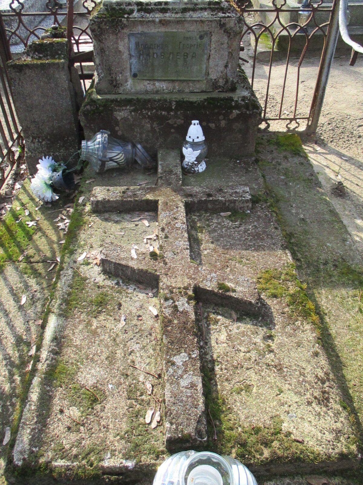 Zdjęcie przedstawia prawosławny nagrobek. W górnej części znajduje się płyta z informacjami na temat zmarłego. Napisy są nieczytelne. Poniżej, widoczny jest wyrzeźbiony krzyż prawosławny. Wokół niego stoją, a także leżą znicze z białymi elementami oraz białe, sztuczne kwiaty. Grób ogrodzony jest metalowym płotem. Zdjęcie zostało wykonane w świetle dziennym.