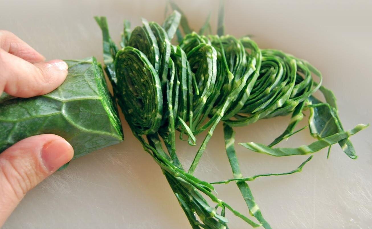Zdjęcie przedstawia listki bazylii pokrojone techniką chiffonade. Na pierwszym planie, po lewej stronie, widoczna jest dłoń trzymająca rulon listków bazylii. Obok pokrojone na cienkie paseczki listki bazylii.