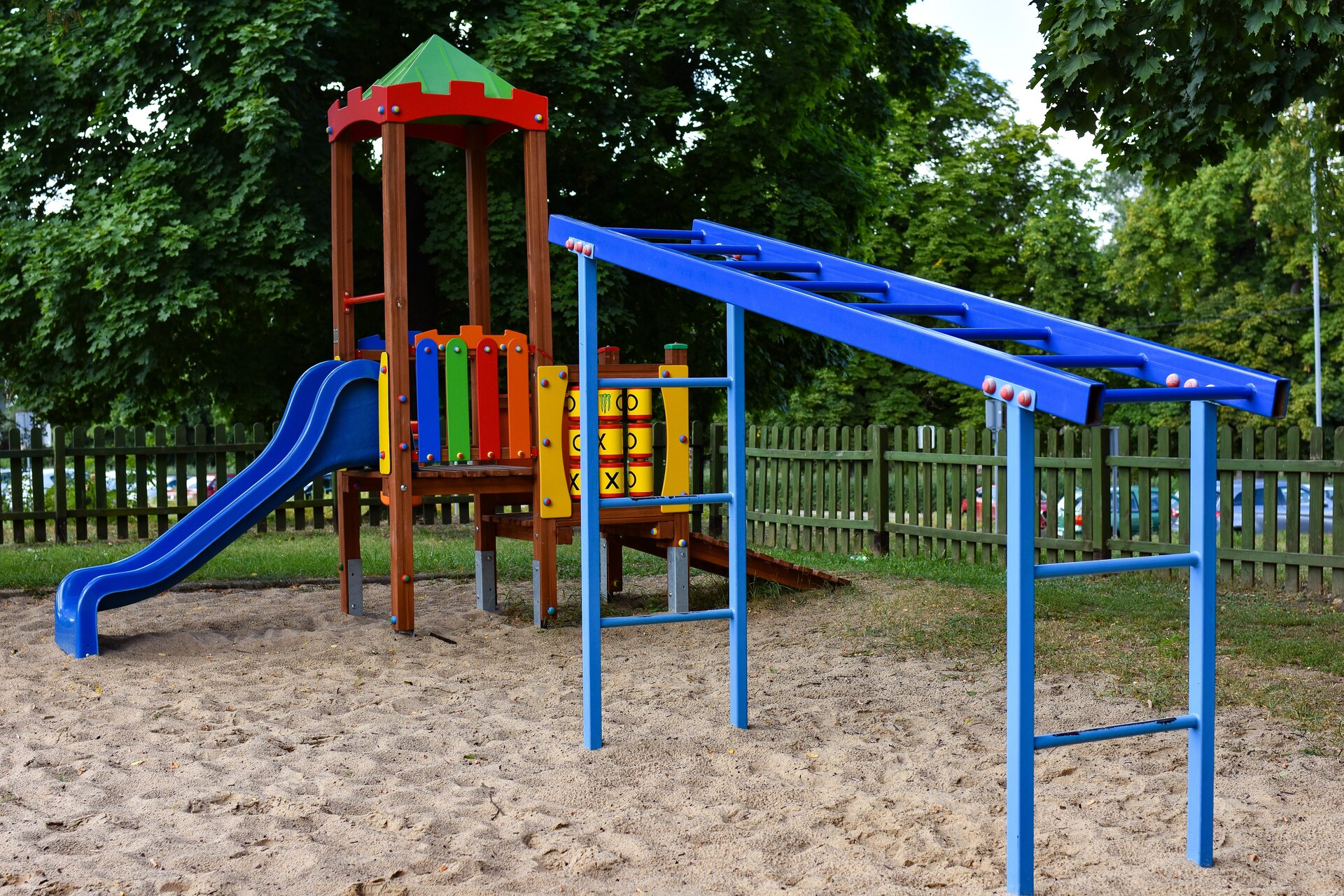 Fotografia przestawia plac zabaw. Wśród atrakcji placu zabaw znajdują się niebieska drabinka, domek ze zjeżdżalnią oraz gra w kółko i krzyżyk. Wyposażenie placu zabaw zbudowane jest z kolorowych elementów. Powierzchnia placu zabaw pokryta jest piaskiem. W tle zdjęcia widoczny jest drewniany płot oraz drzewa.