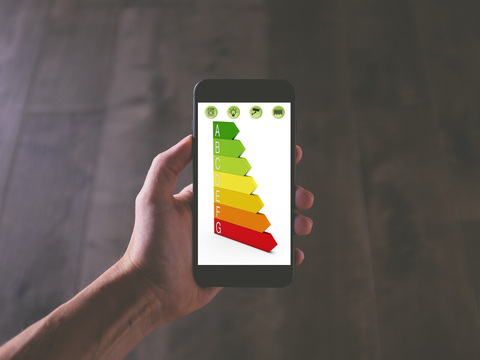 Ilustracja przedstawia lewą dłoń, która trzyma telefon komórkowy. Na ekranie wyświetlono skalę efektywności energetycznej dotyczącą sprzętu AGD. Skala oznaczona jest kolorowymi strzałkami a każda opisana jest  literami od A do G. Transkrypcja znajduje się poniżej
