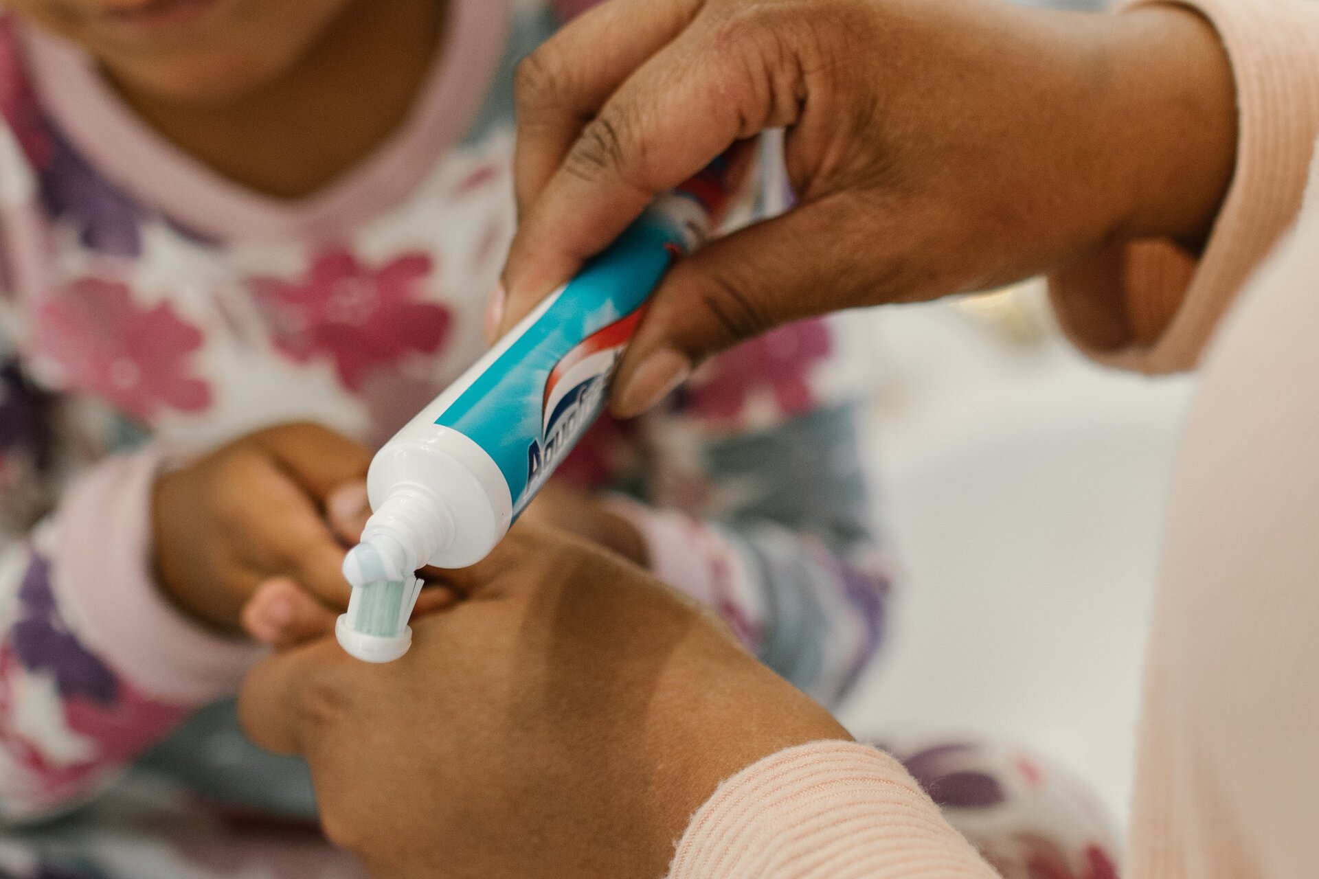 Zdjęcie przedstawia dziecko trzymające szczoteczkę do mycia zębów oraz dorosłą osobę nakładającą pastę do zębów na szczoteczkę dziecka.