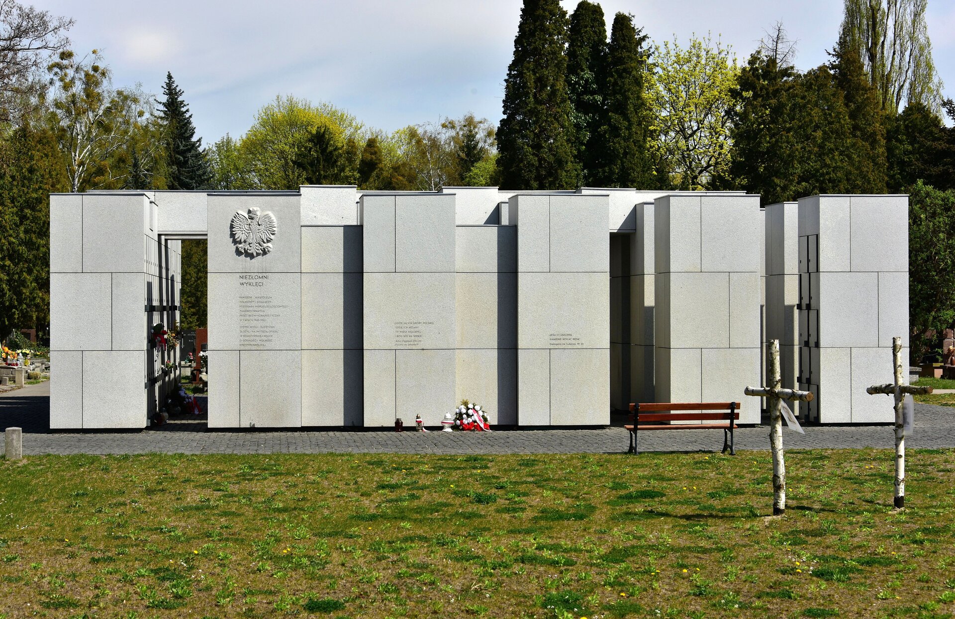 Zdjęcie pokazuje elewację kamiennego pomnika z wizerunkiem orła białego i tablicą informacyjną, pod którą jest wieniec i znicze. Pomnik składa się z sześciu bloków‑filarów, z których skrajne są oddzielone od pozostałych, a środkowe tworzą ścianę z tablicami informacyjnymi. Przed elewacją pomnika przy chodniku stoi drewniana ławka. Na pierwszym planie jest trawnik. Po jego prawej stronie na zdjęciu stoją dwa brzozowe krzyże. W tyle zdjęcia za pomnikiem rosną drzewa. 