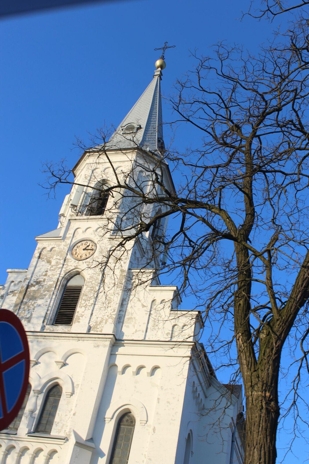 Zdjęcie przedstawia białą, wysoką wieżę, której dach jest szary i skośny. Na czubku wieży znajduje się krzyż. Zdjęcie ukazuje przód kościoła. Przed budynkiem rośnie bezlistne drzewo. Niebo jest błękitne, bezchmurne. Zdjęcie zostało wykonane w świetle dziennym.