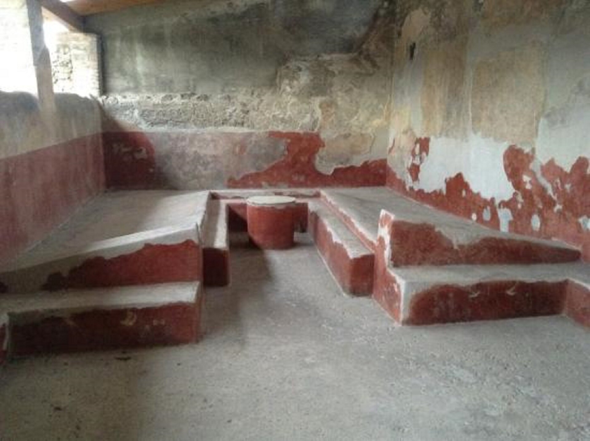 Fotografia nieznanego autorstwa przedstawia murowane triclinium w Pompejach. Na fotografii obecne są trzy ławki wykonane z cegły. Triclinum znajduje się w zniszczonym pomieszczeniu, w którym z części ścian odchodzi czerwona farba. 