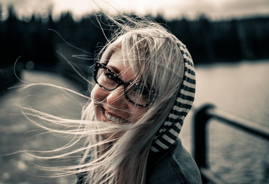 Fotografia kolorowa przedstawia kobietę w okularach z ciemną oprawką, na którą wieje wiatr. Rozrzucone włosy wystają spod kaptura w poziome paski, kobieta uśmiecha się do kamery, stoi na moście, bądź też kładce przy brzegu zbiornika wodnego widocznego w tle.