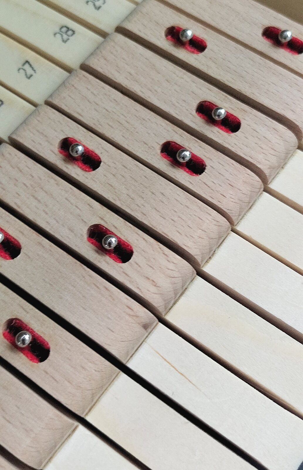 Zdjęcie przedstawia klawisze wykonane z drewna świerkowego oraz nakładki środkowe wykonane z drewna bukowego. Widoczny jest rząd jasnobeżowych klawiszy, na które nałożone są jasnobrązowe prostokątne nakładki, na których w nieregularnym układzie widoczne są elipsy z czerwonymi krawędziami na boku i czarnym paskiem w środku, wewnątrz których widoczne są srebrne kulki. Powyżej widoczny jest rząd z częściowo widocznymi jasnymi klawiszami z numerami.
