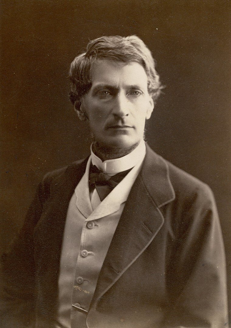 Zdjęcie przedstawia mężczyznę. Ma krótkie, zaczesane na bok włosy oraz lekki zarost na dole brody. Ubrany jest w garnitur.