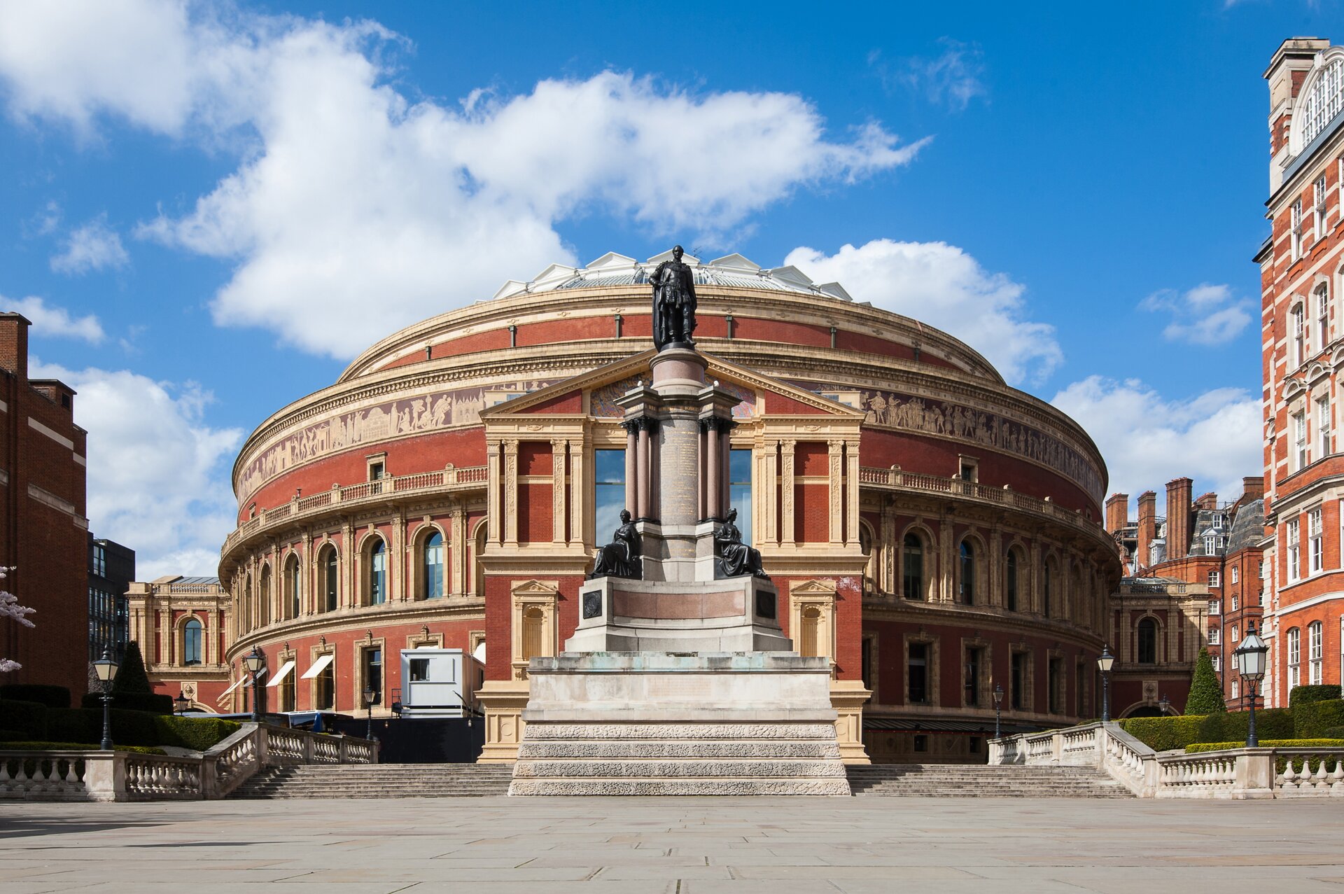 Zdjęcie przedstawia budynek teatru Royal Albert Hall. Na pierwszym planie przed budynkiem znajduje się posąg mężczyzny, który został umieszczony na wysokiej kolumnie. Poniżej, u podstawy kolumny znajdują się dwa inne posągi przedstawiające siedzące kobiety. Budynek ma owalny kształt, jest koloru czerwono‑beżowego. Na fasadzie budynku znajdują się obrazy, przedstawiające różne sceny. Na dachu budynku znajduje się szklana kopuła. Do budynku, po prawej oraz po lewej stronie pomnika prowadzą schody. 