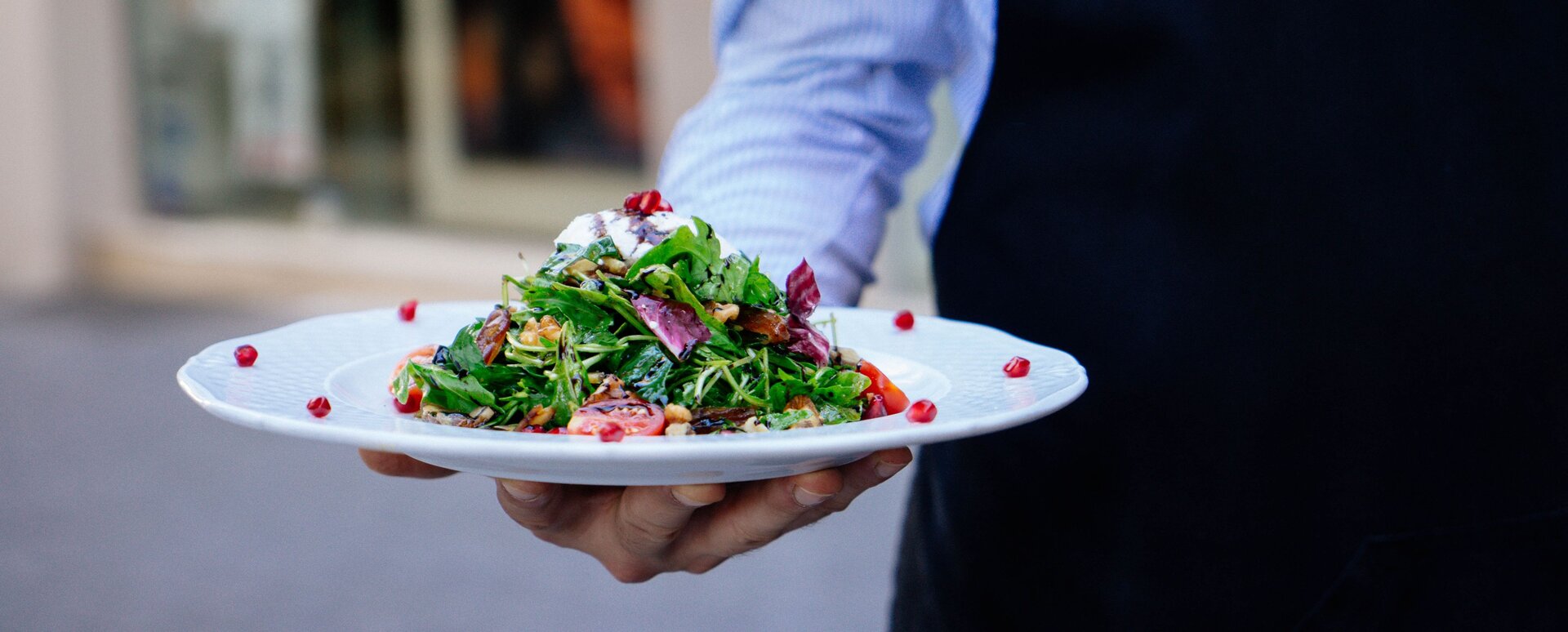 Ilustracja przedstawia talerz z sałatką trzymany przez kelnera.
