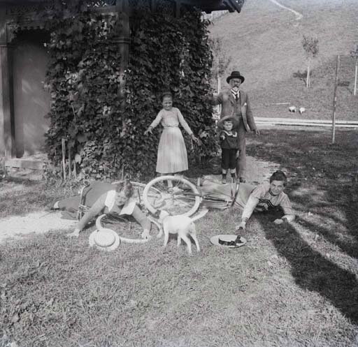 Na zdjęciu dwie leżące na ziemi kobiety w sukniach obok połamanego roweru, przed nimi stojący tyłem piesek. Za rowerem starszy mężczyzna z wąsami, w kapeluszu na głowie, chłopczyk także w kapeluszu i starsza dziewczynka w sukience. Za nimi budynek z pnącym się po ścianie bluszczem.