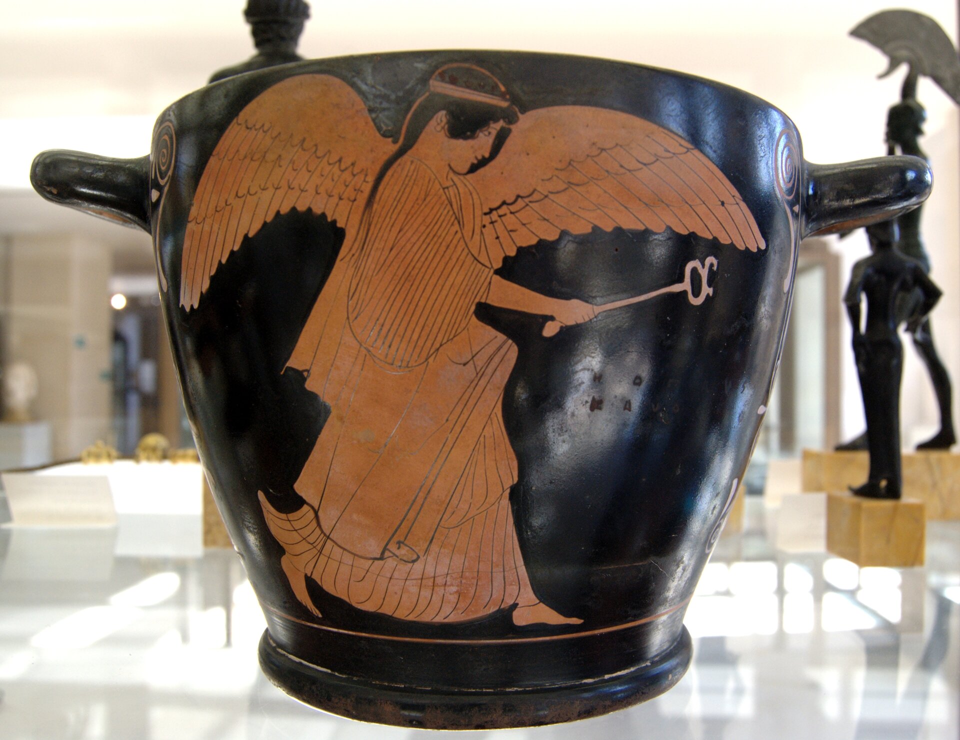 Zdjęcie przedstawia attycką wazę z uchami z obu stron  zwaną skyfosem. Na niej namalowano boginię Iris, kobieta ma długie włosy, potężne skrzydła oraz długą, fałdowaną szatę. W dłoni trzyma długi pręt zakończony okręgiem. Tło jest czarne, a postać rdzawa. W tle widać inne eksponaty muzealne.