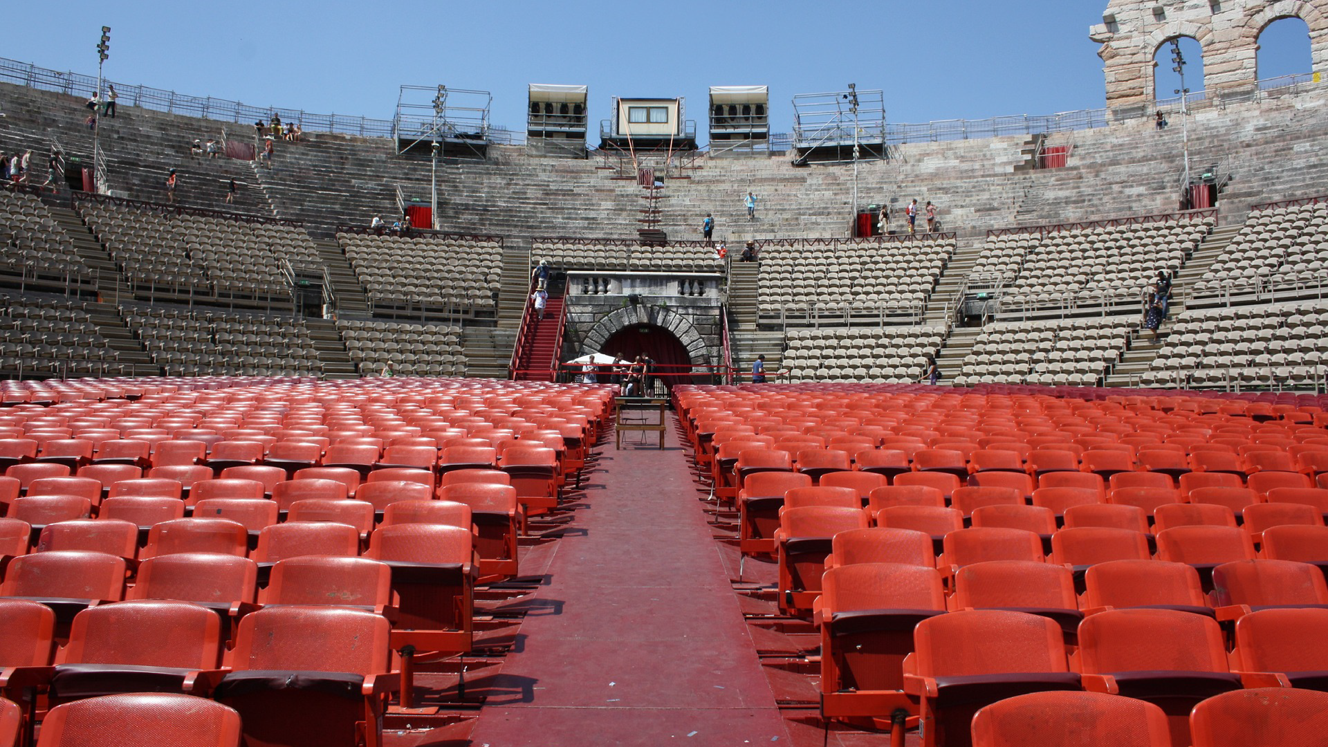 Zdjęcie przedstawia wnętrze amfiteatru w Weronie. Teatr znajduje się pod gołym niebem. Na zdjęciu na pierwszym planie widoczne są rzędy krzeseł. Na drugim planie widoczne są trybuny amfiteatru.