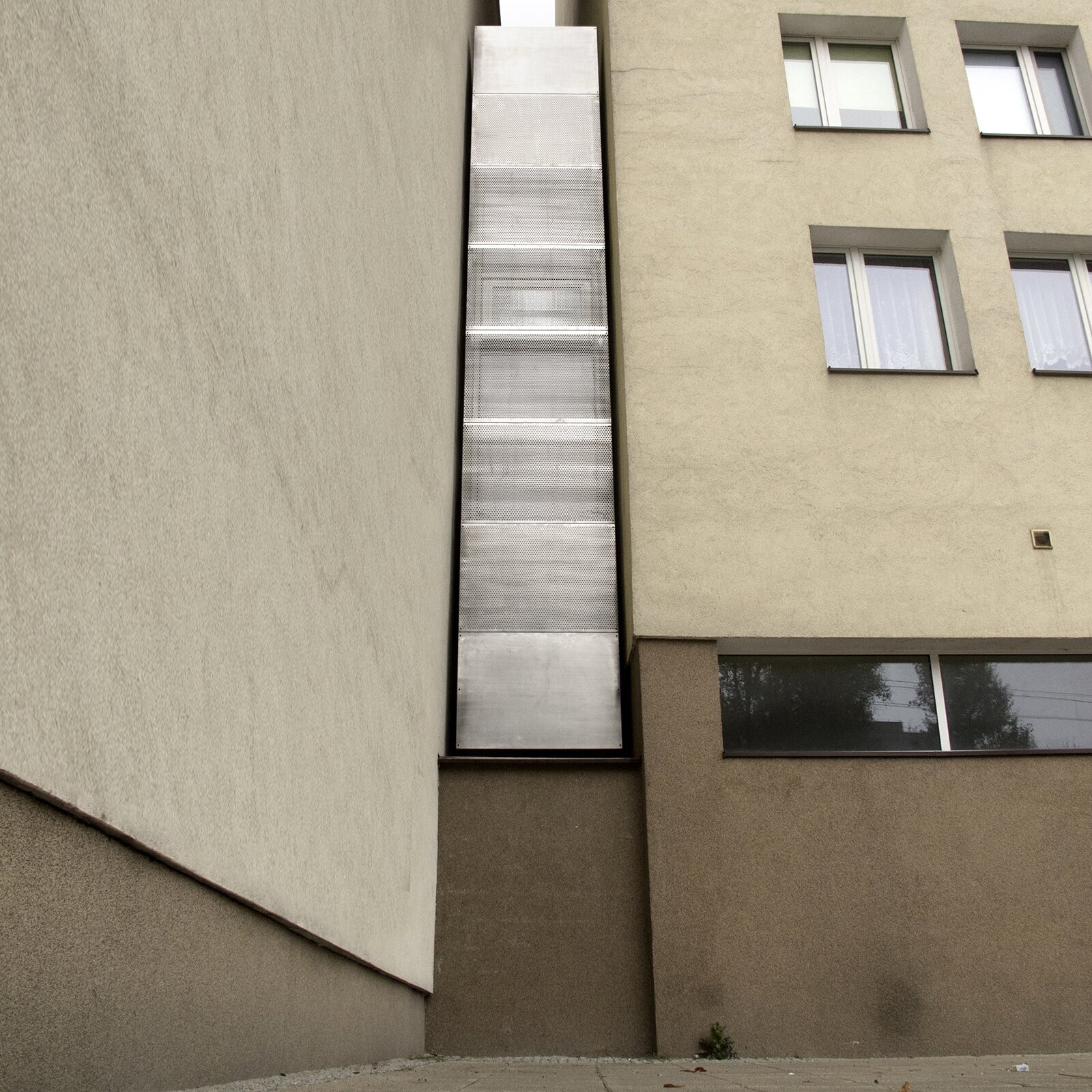 Na zdjęciu widzimy zbliżenie na dwie ściany wysokiego bloku, połączone w literę el. Po prawej stronie widać okna, a przy połączeniu ścian znajduje się podłużna, prostokątna konstrukcja z metalu, łącząca piętra budowli. 