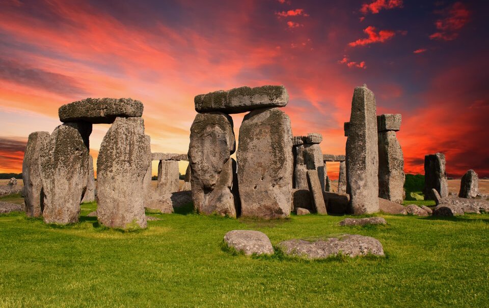 Fotografia przedstawia Stonehenge. Wielkie kamienie ułożone pionowo z poprzecznymi znacznie mniejszymi elementami. Wszystkie owe struktury są ułożone w koncentryczne kręgi, z których część rozpadła się już i porosła częściowo trawą.
