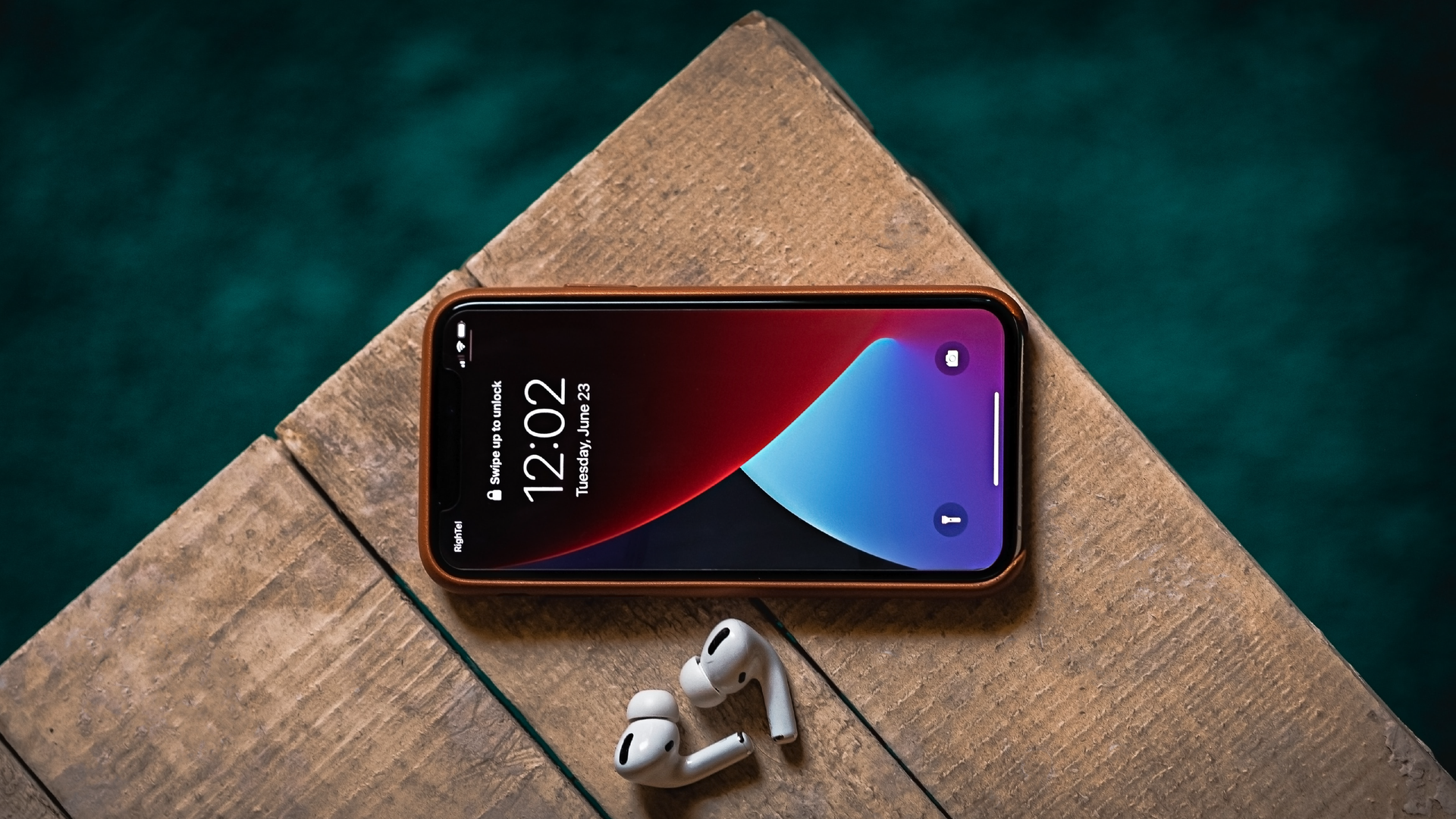 Zdjęcie przedstawia telefon komórkowy, który leży na drewnianym pomoście nad wodą. Obok telefonu leżą bezprzewodowe słuchawki douszne.