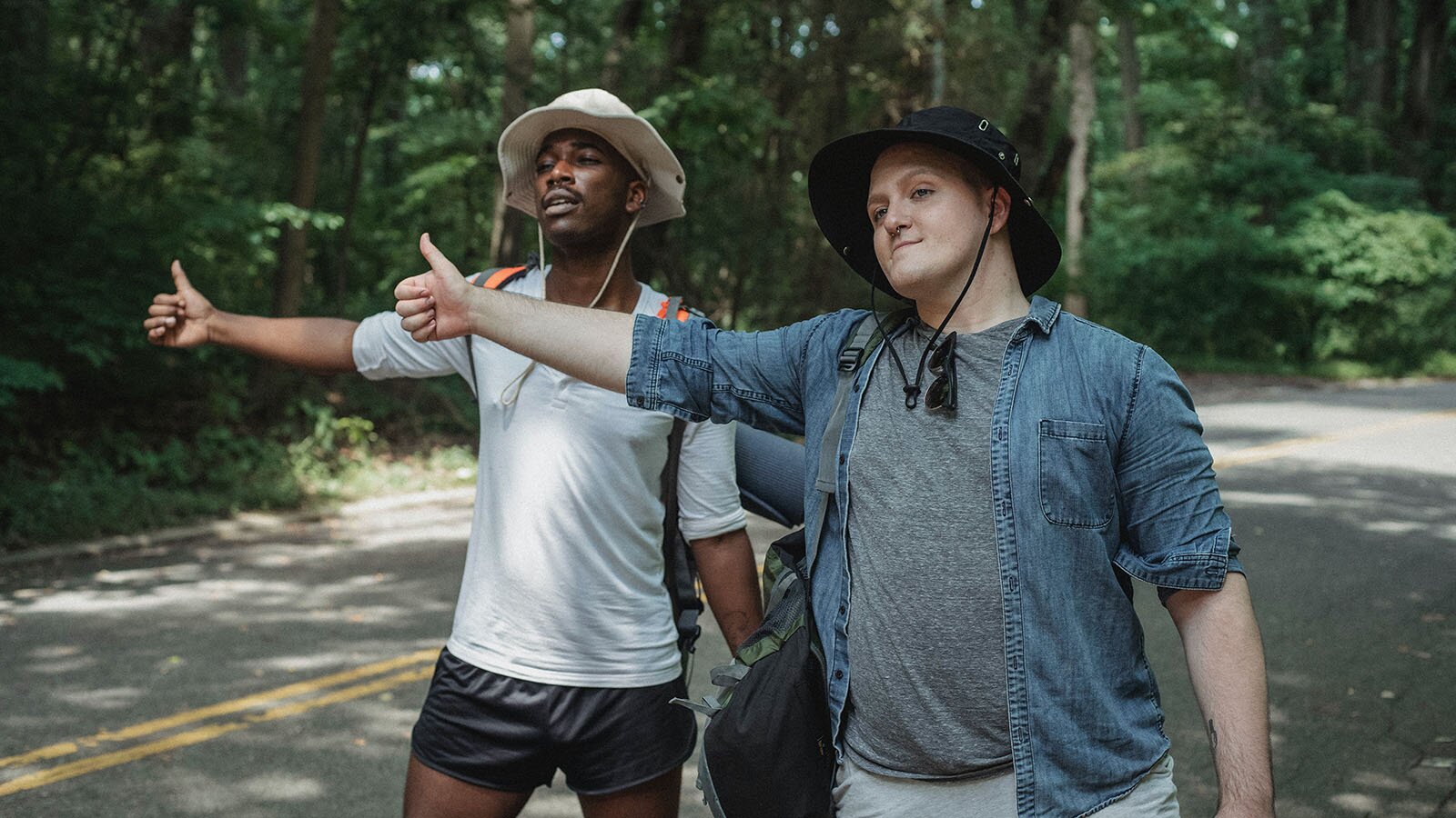 Zdjęcie przedstawia dwóch młodych mężczyzn stojących na drodze w lesie. Wyciągają ręce z kciukiem do góry chcąc złapać stopa. 