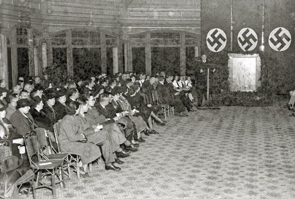 Zdjęcie przedstawia kobiety i mężczyzn, którzy siedzą na krzesłach po lewej stronie pomieszczenia. Po prawej stronie stoi mównica oraz wiszą trzy sztandary z hitlerowskimi swastykami.