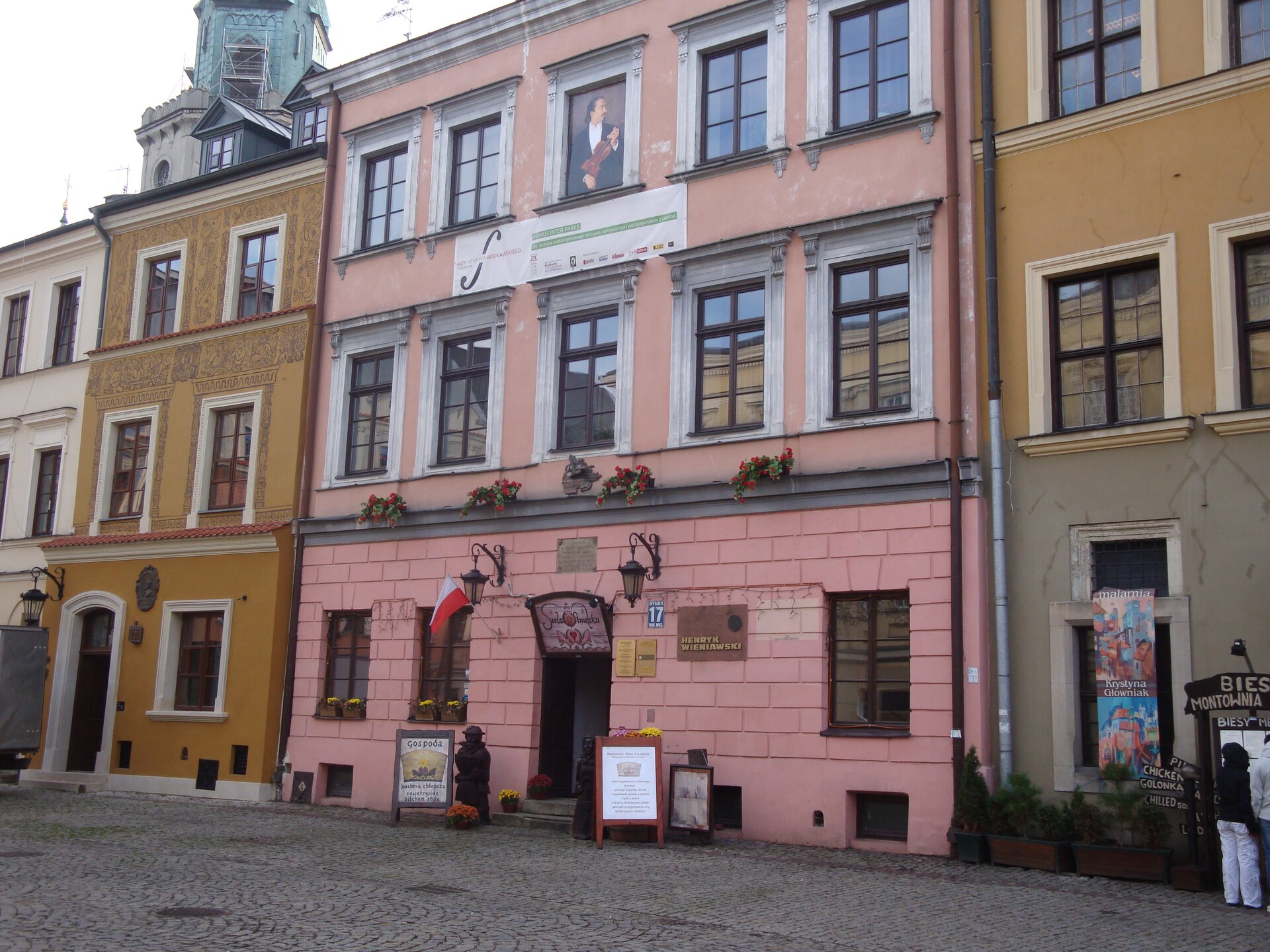 Ilustracja przedstawia domy na lubelskiej starówce. W centrum znajduje się dom, w którym urodził się Henryk Wieniawski. Fasada tej kamienicy jest pomalowana na różowo. Budynek ma dwa piętra. Na pierwszym piętrze 5 okien, na drugim piętrze zamiast środkowego okna umieszczono portret kompozytora.