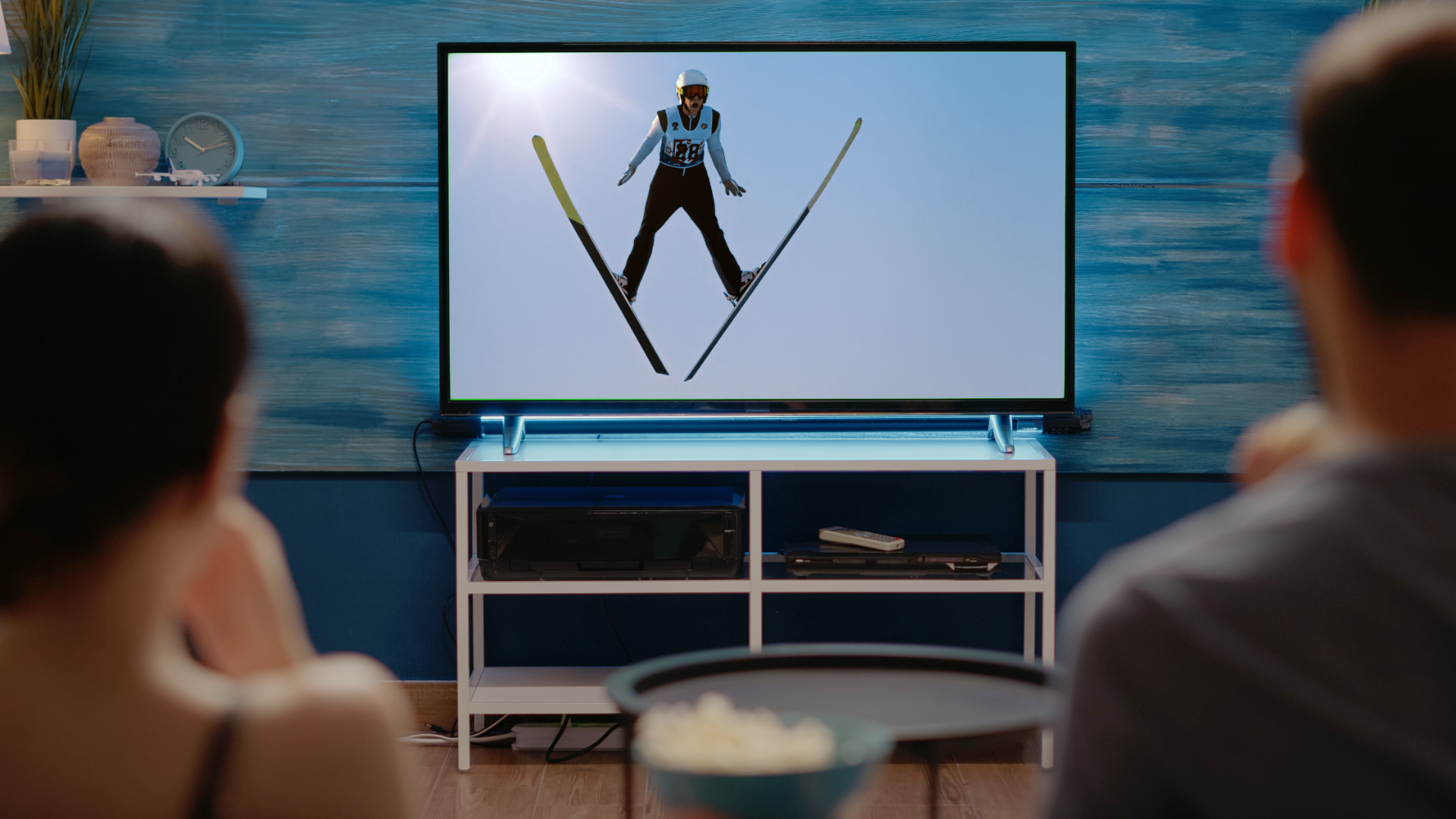 W środkowej części zdjęcia znajduje się telewizor, na którym widoczny jest skoczek  narciarski w czasie skoku. Na pierwszym planie, tyłem do kadru, a przodem do telewizora siedzą kobieta i mężczyzna.